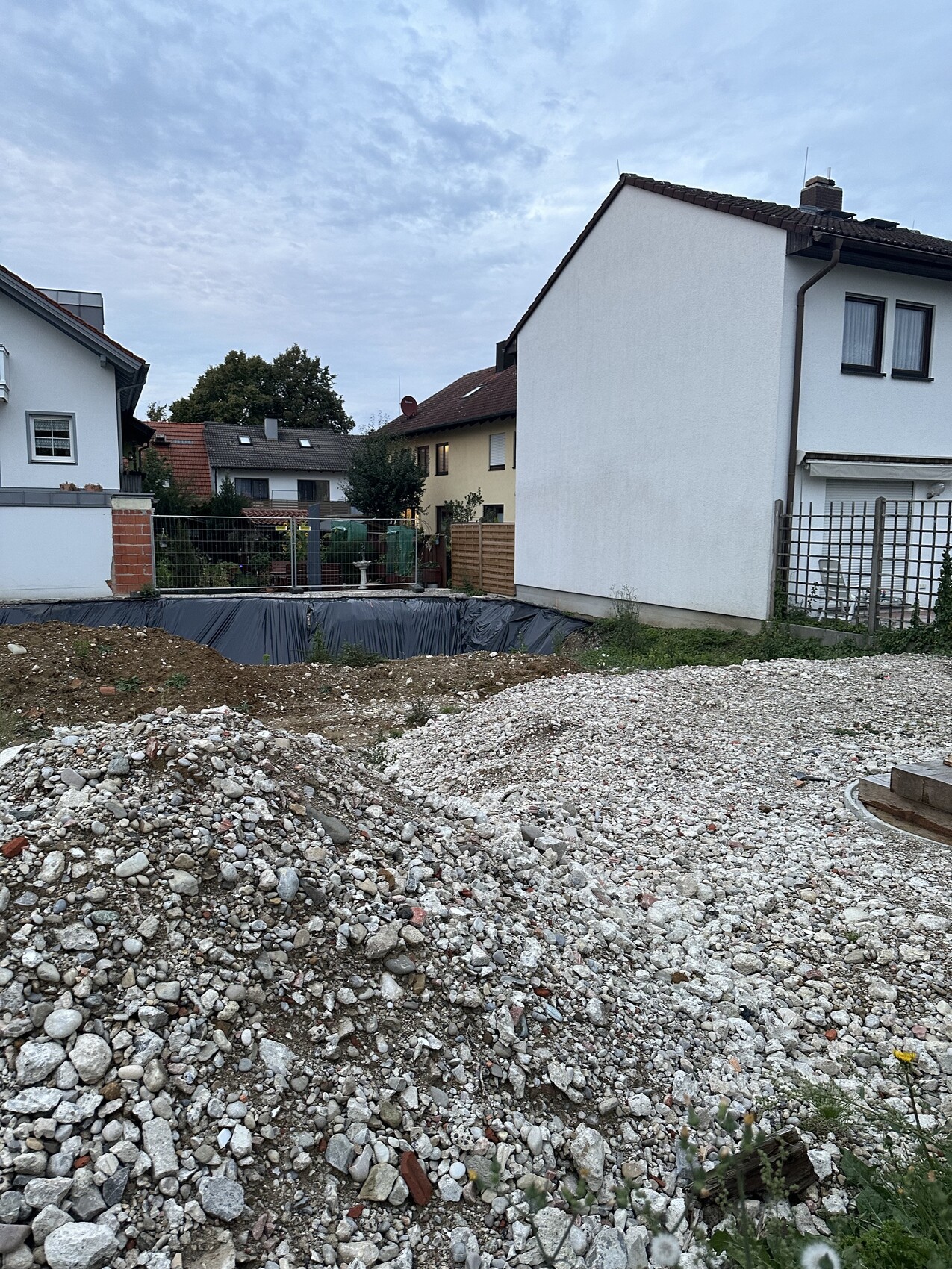 Large excavation pit in a residential area, in front gravel and rubble piles, adjacent houses.