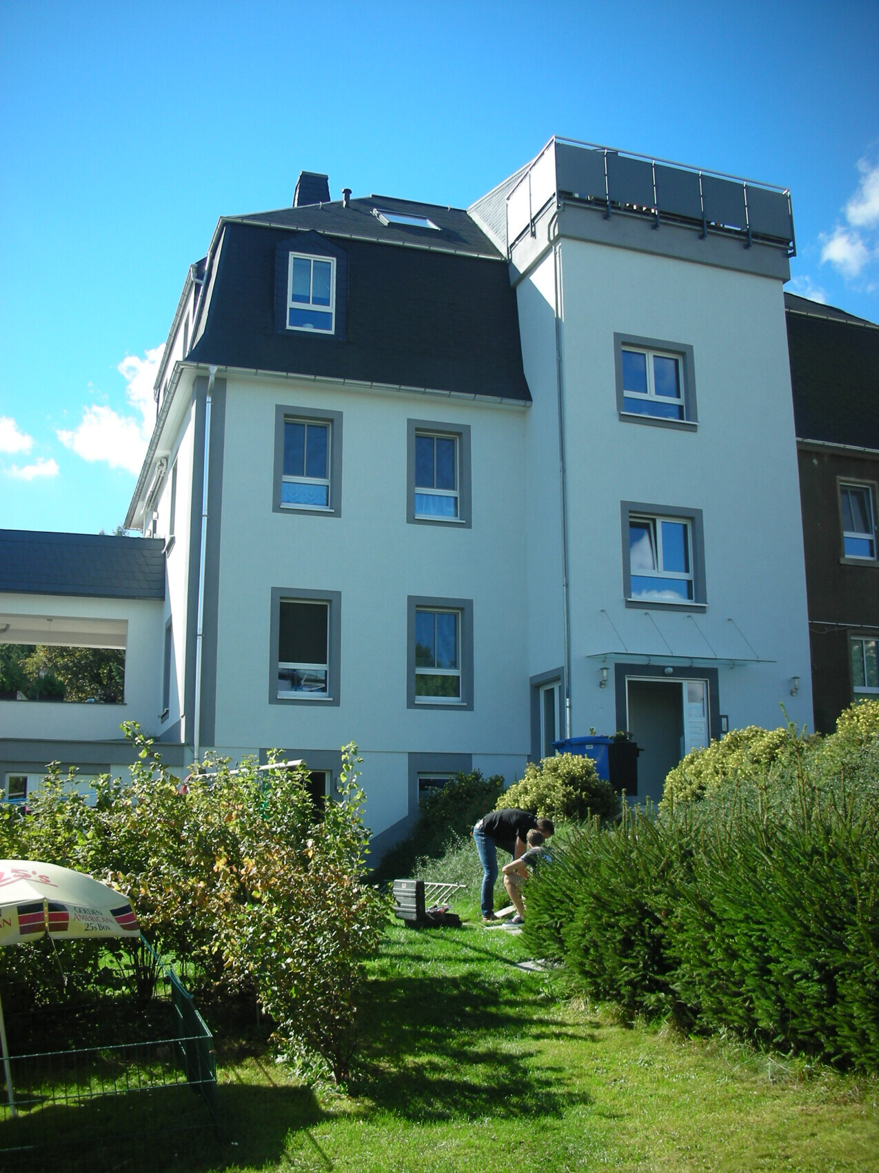 Bright multi-family house with dark roof, garden, and two people gardening in the front yard.