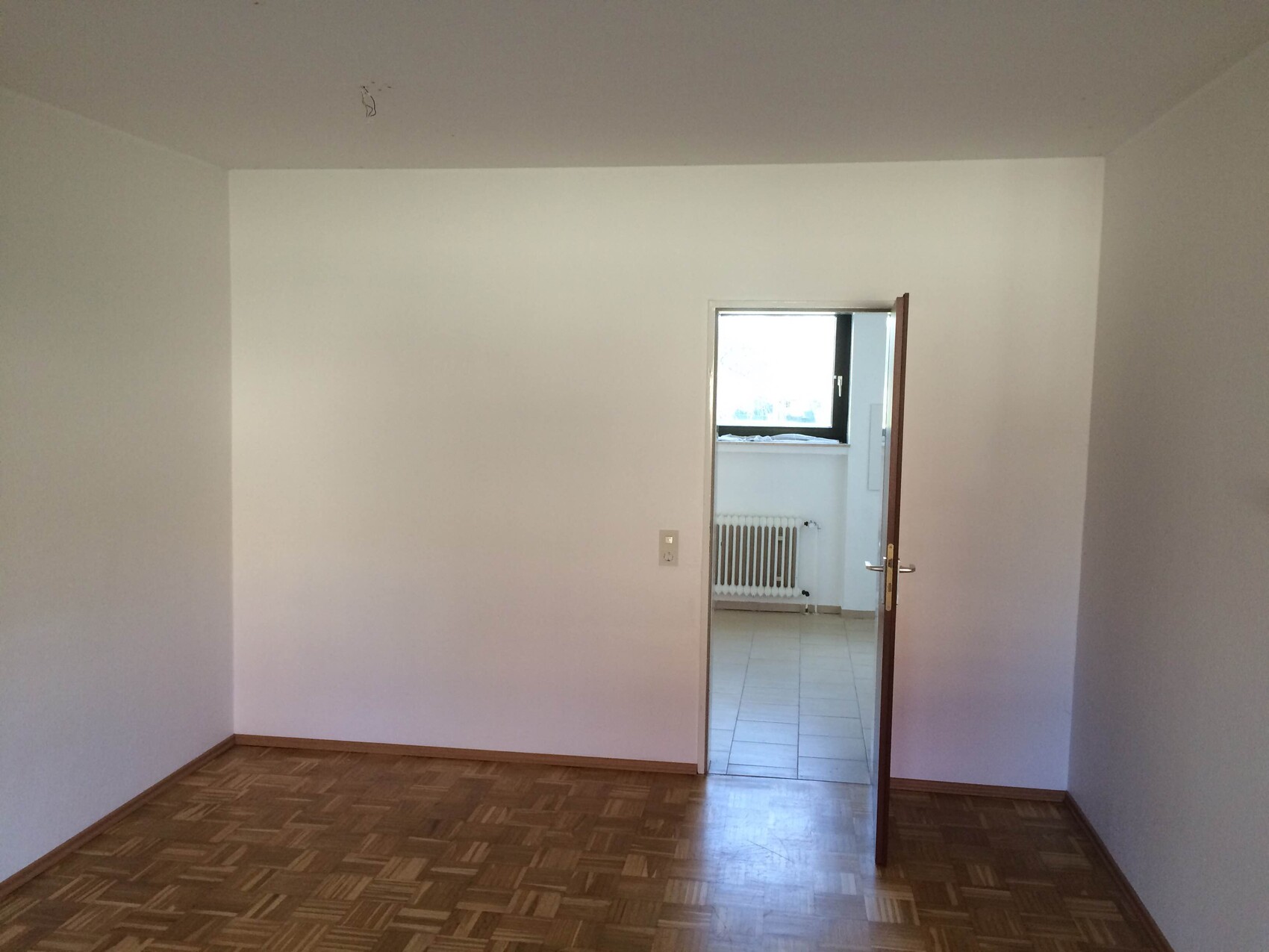 Empty living room with light-colored walls, parquet floor, and open door to the adjacent room