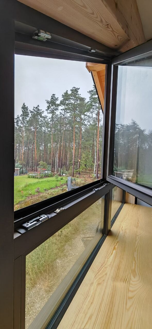 Open window with black metal frame and wooden floor; view of pine forest, meadow, and garden outside.