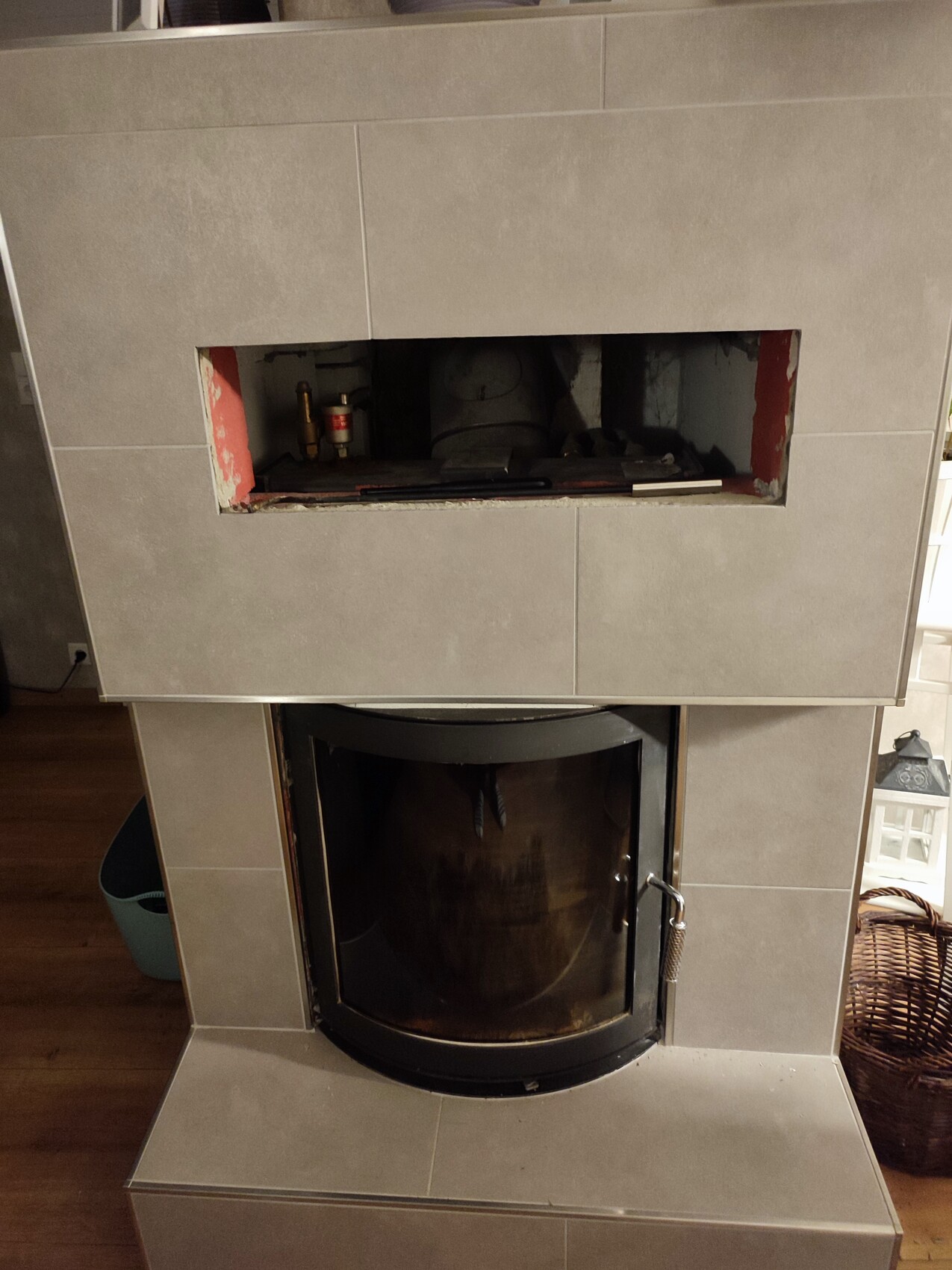 Modern wood stove with round glass door; above it an open compartment with visible pipes.
