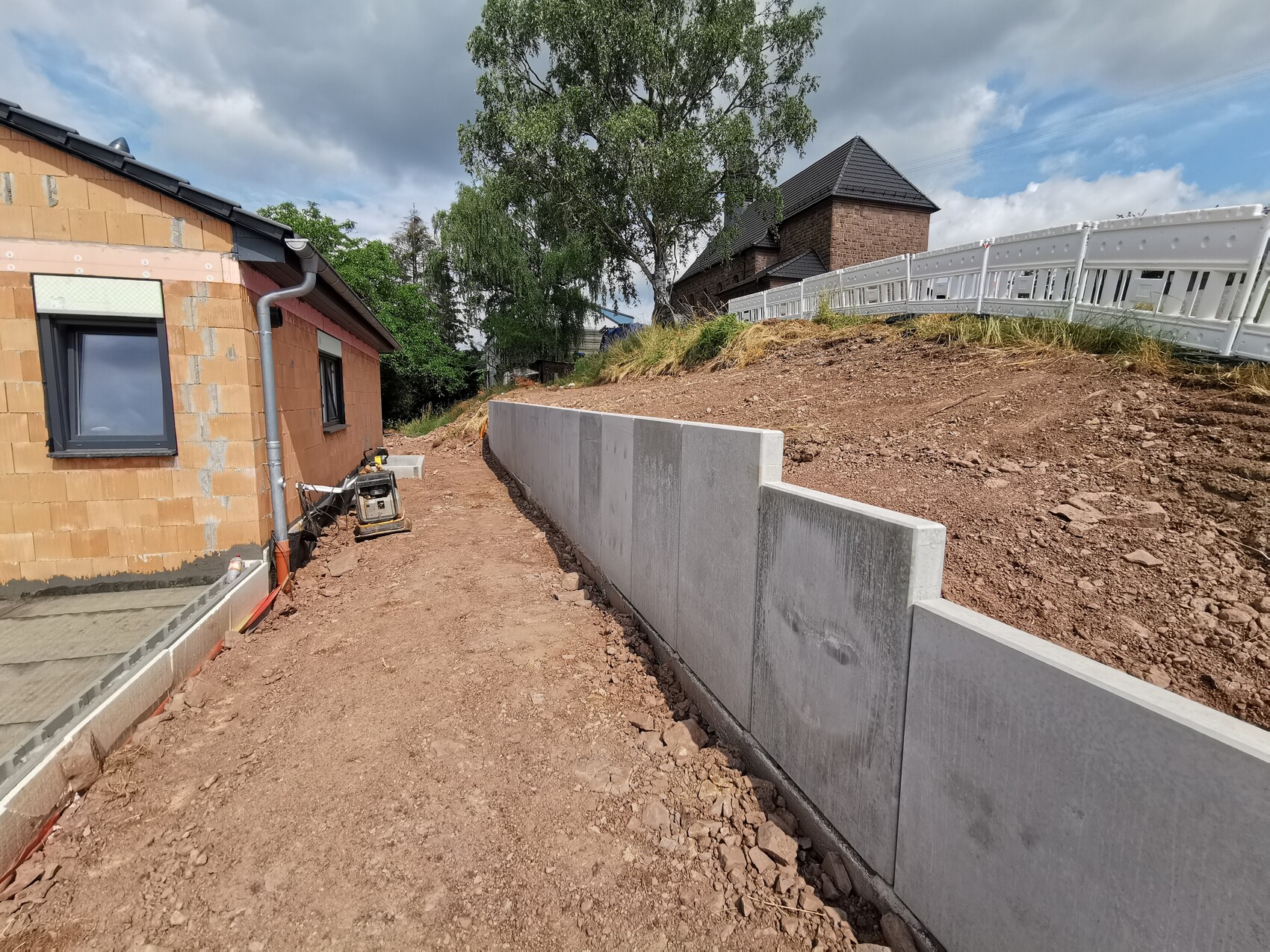 Construction site on a slope: red brick house, concrete wall, construction equipment, and fencing.