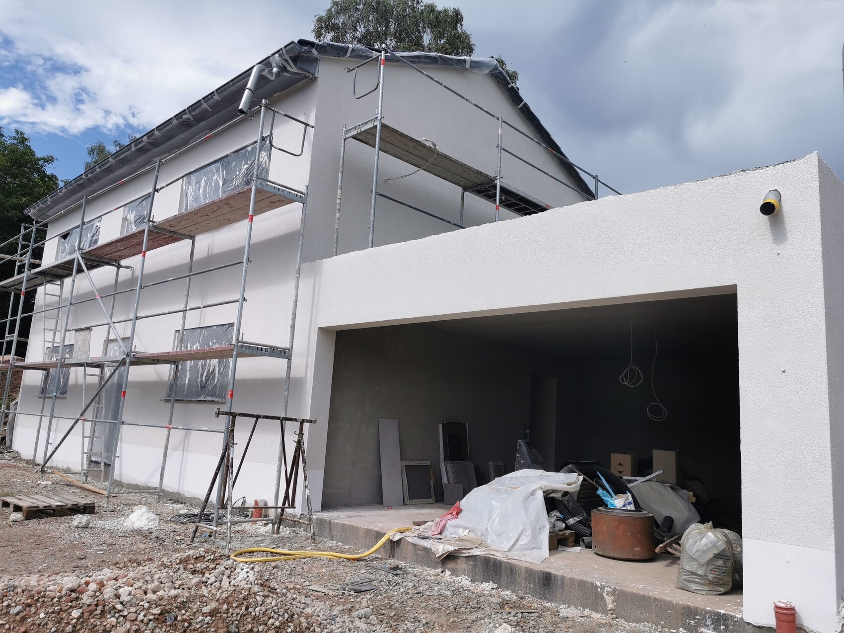 Modern house under construction with scaffolding, white facade, open garage, and building materials.
