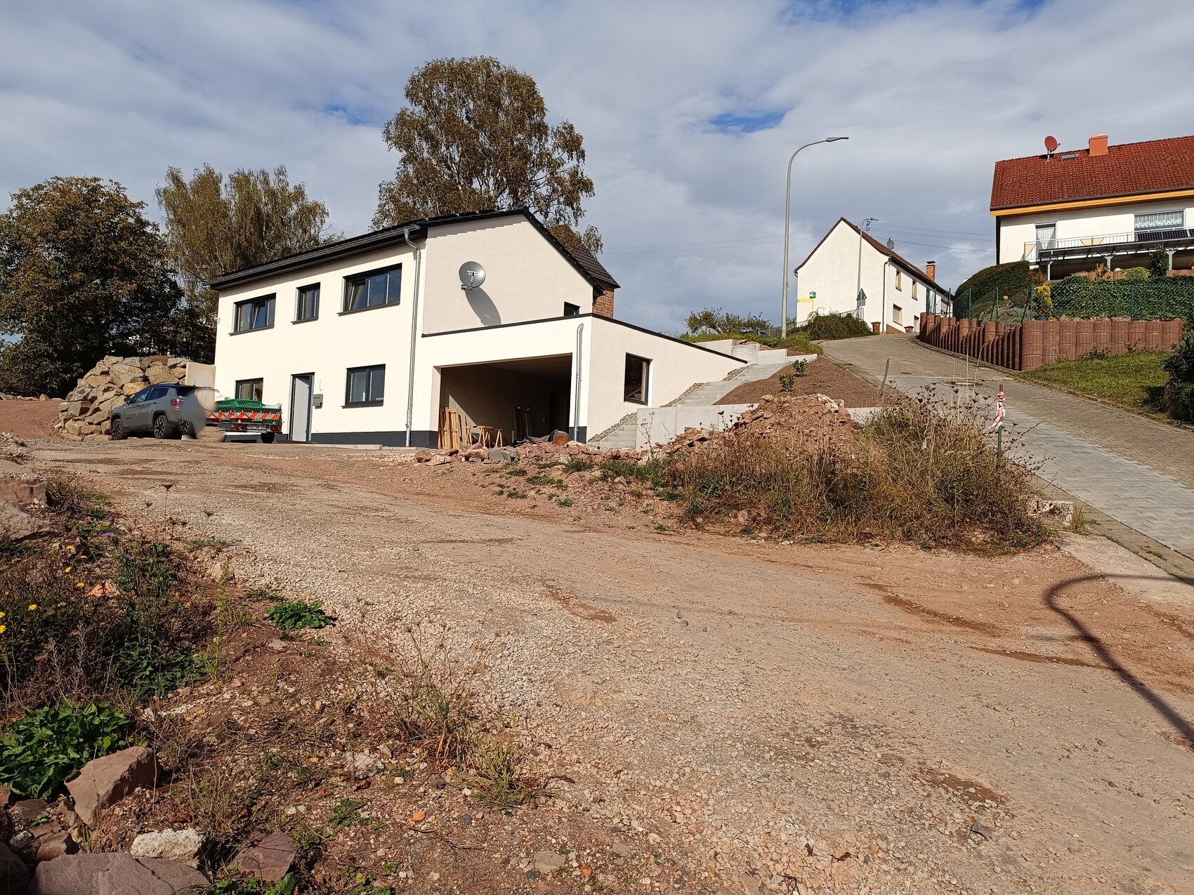 White, two-story new build villa with carport; construction site with gravel, stones, and car.