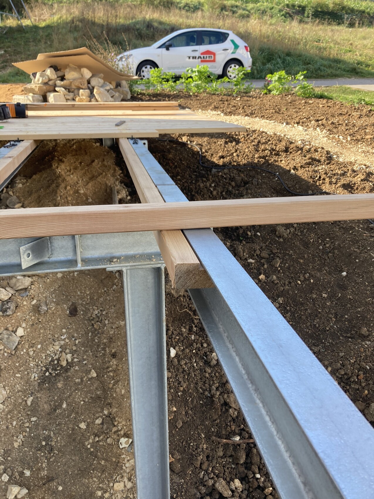 Construction site: steel beams and wooden joists over a trench; stones, tools, car with grape logo behind.