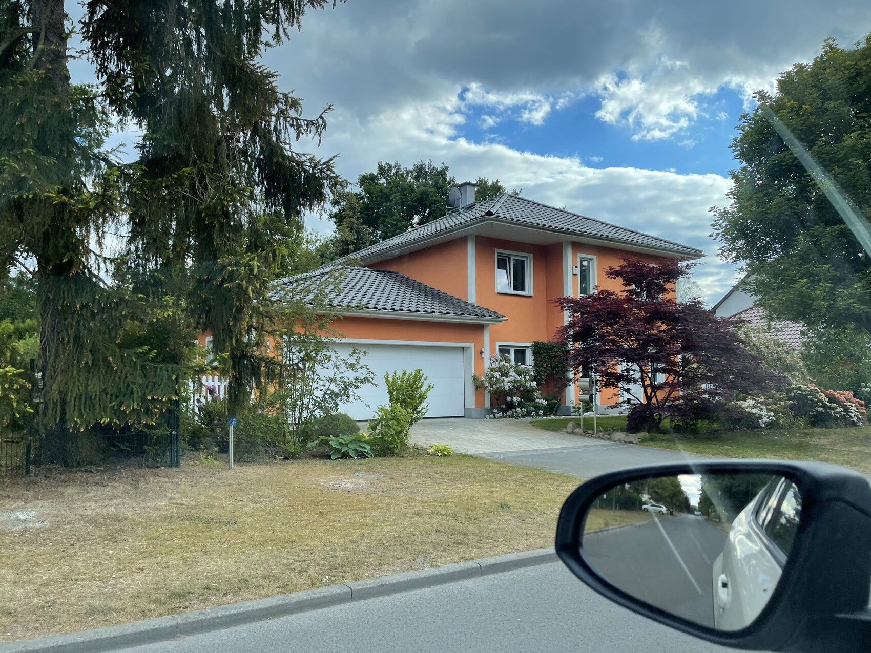 Orange two-story house with garage, garden, and trees; car mirror in the foreground.