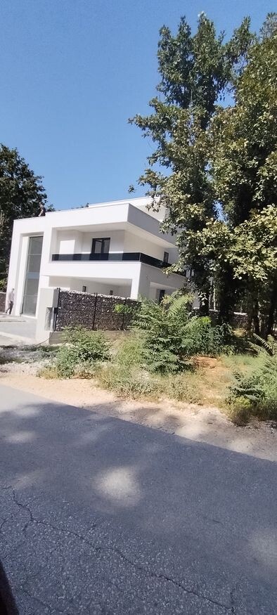 Modern white two-story house with balcony, fence, and large trees on the edge.