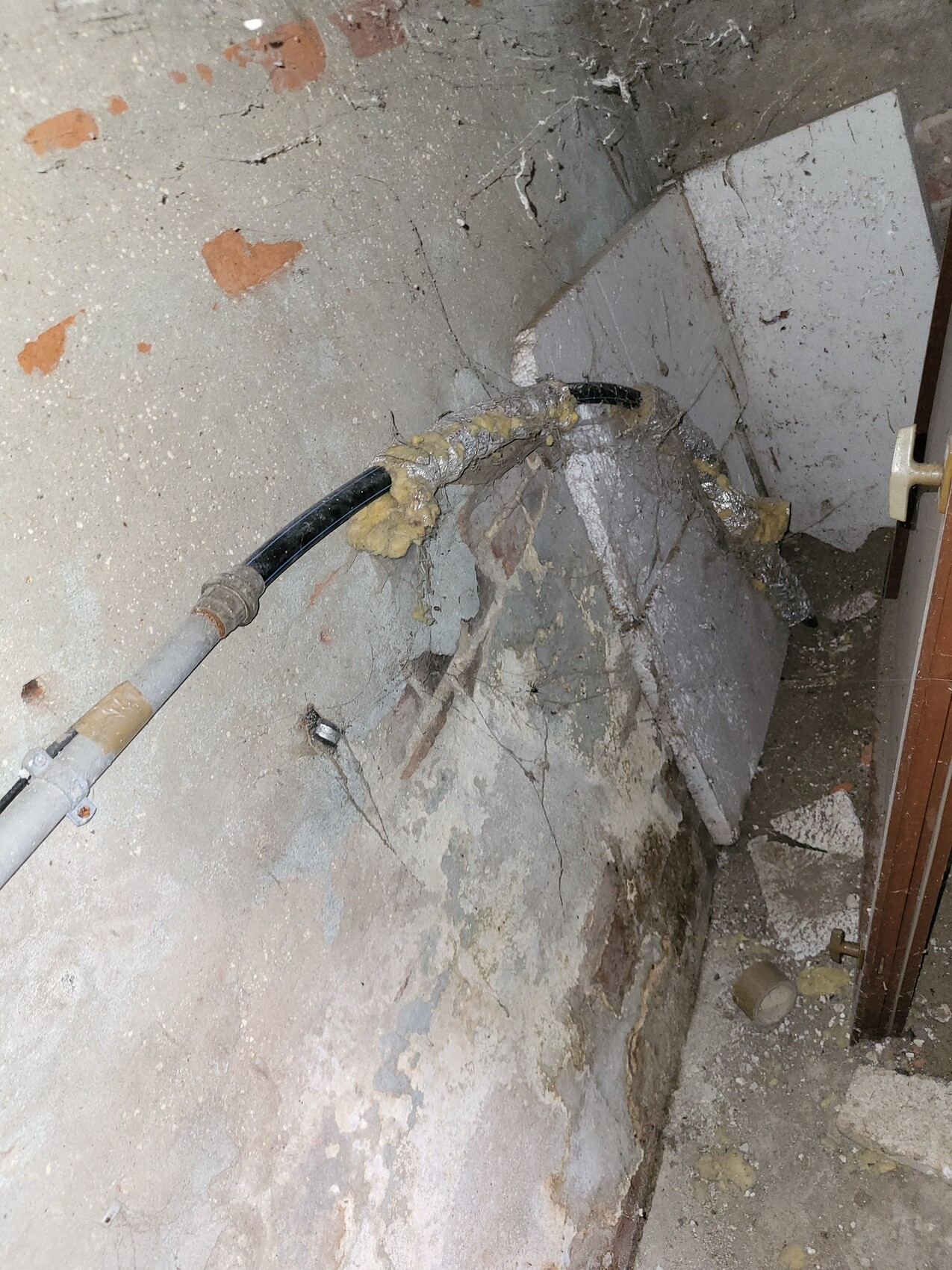 Damp basement wall with piping and crumbling insulation; dust on the floor.