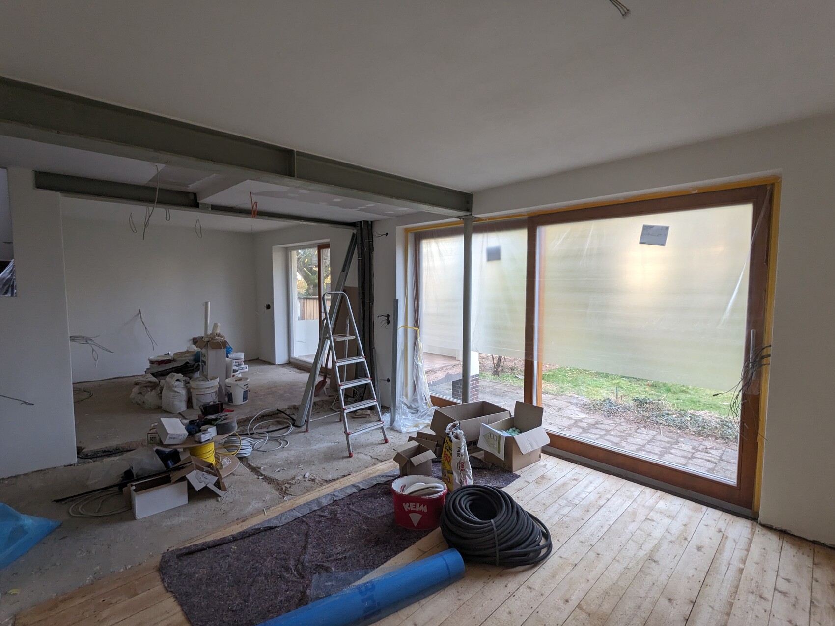 Baustelle im Wohnzimmer: Leiter, Baumaterialien, lose Kabel, große Glasfenster mit Folie.