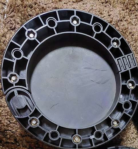 Round black plastic cover with bolt ring, lying on a brown carpet.