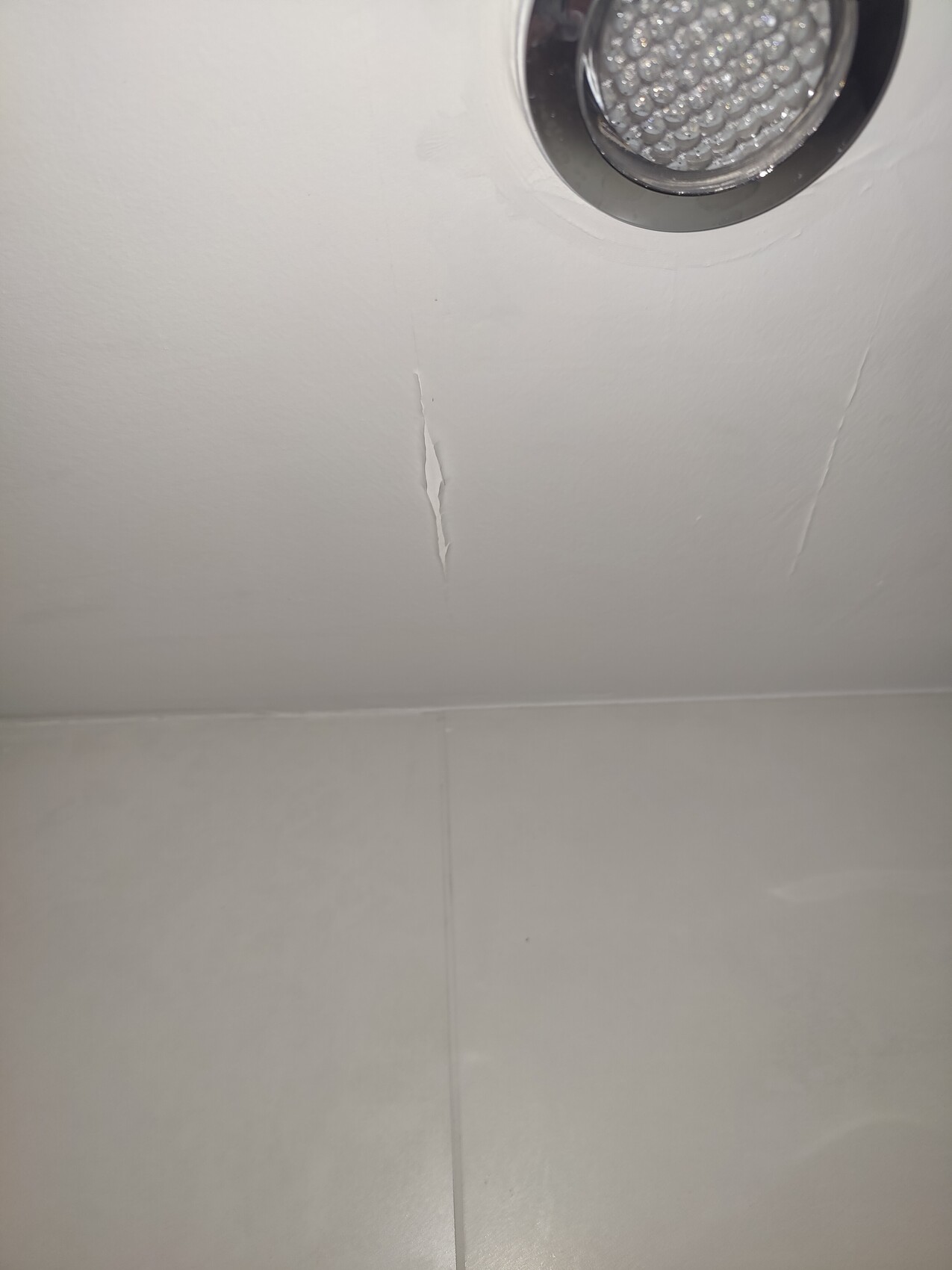 Round LED ceiling spotlight on white ceiling; crack/spalling in the ceiling surface below.