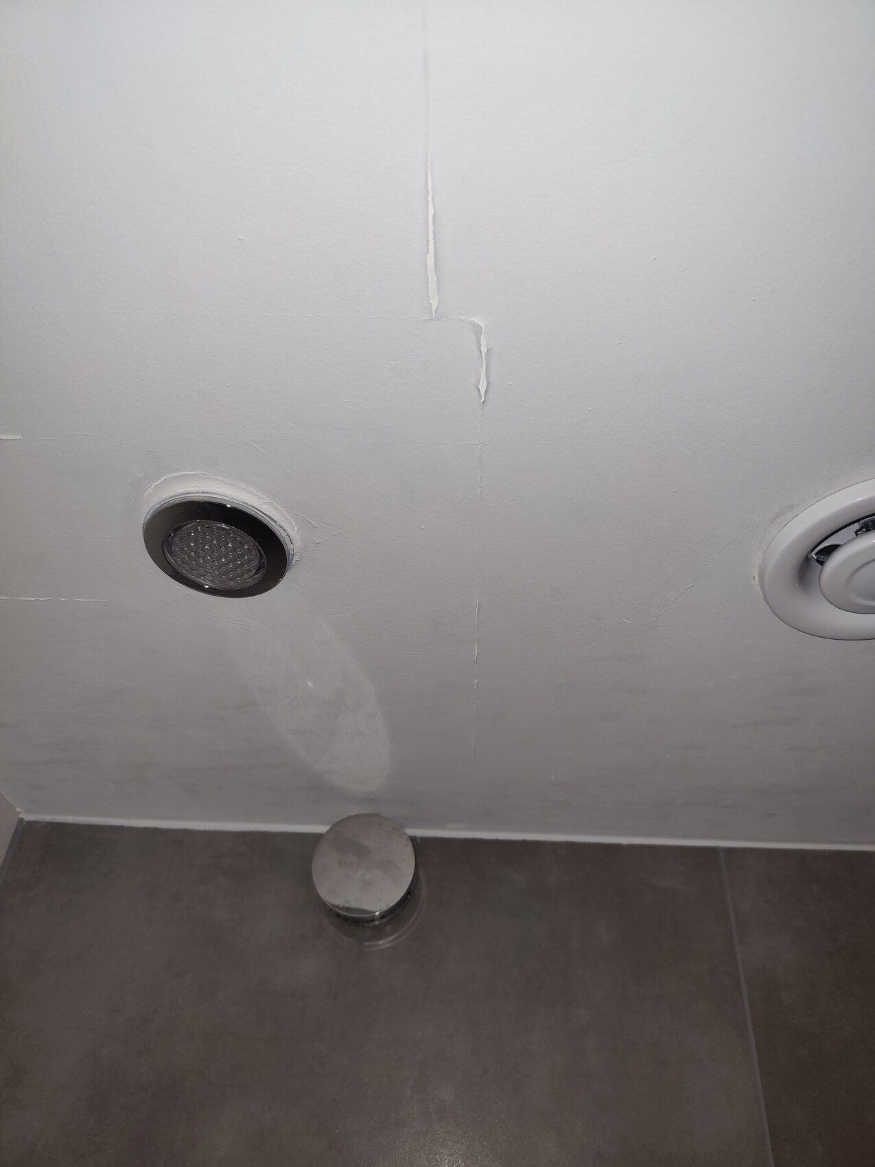 White bathroom wall with cracks; left round metal opening, floor with cover.