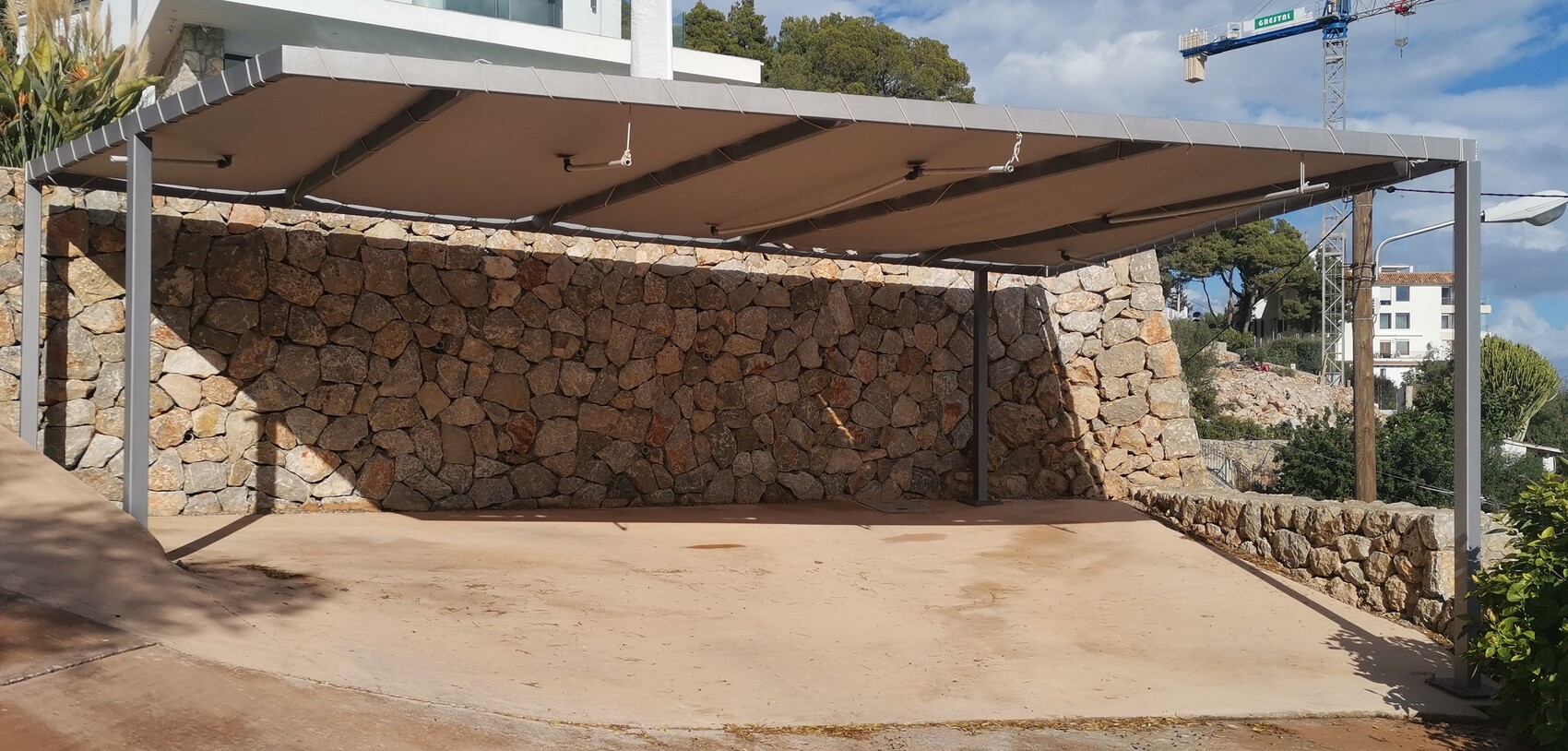 Steel carport with brown fabric roof in front of a stone wall; construction crane and houses in the background.