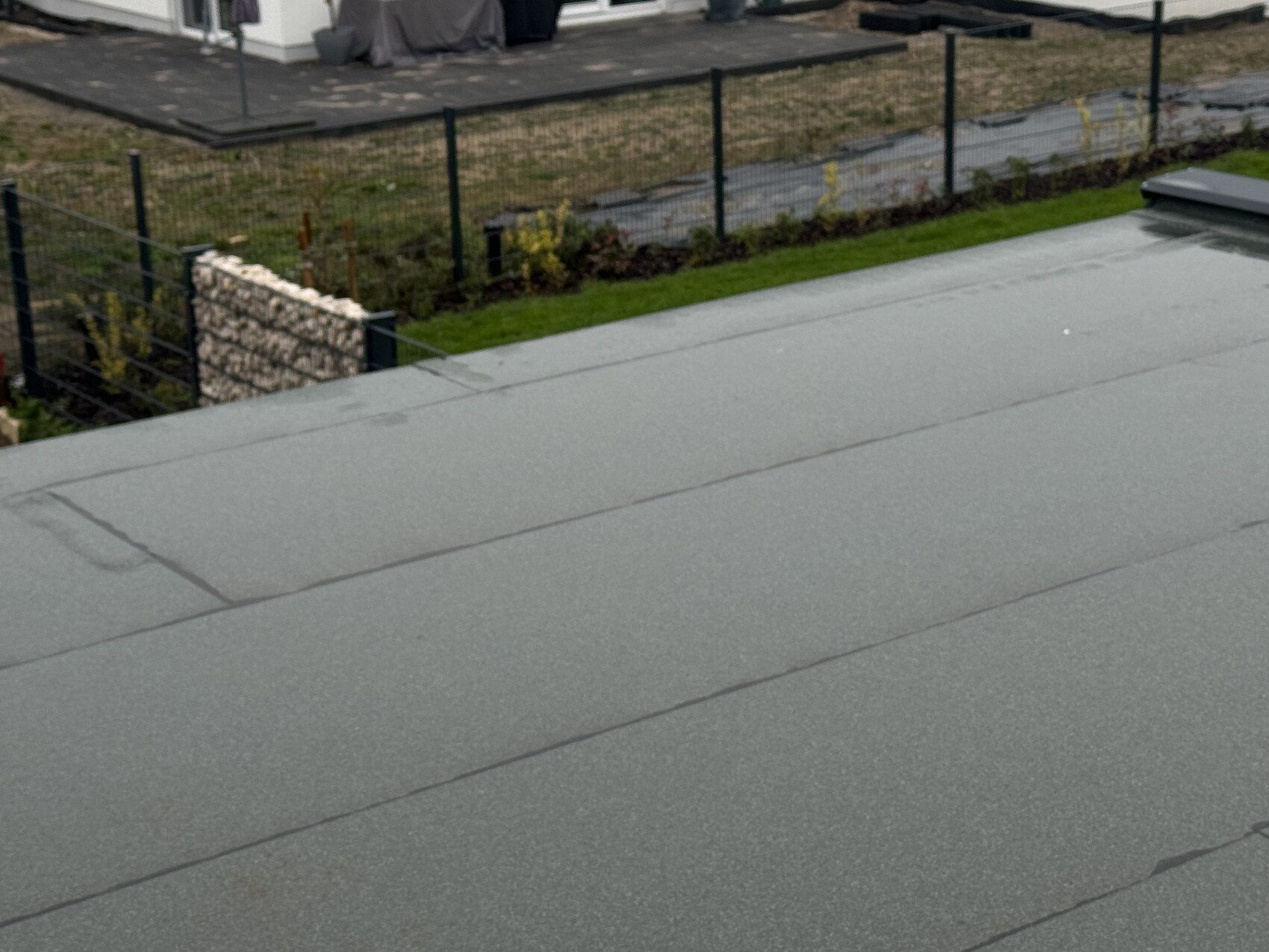 On a gray flat roof, behind it fence, garden with stone wall and greenery.