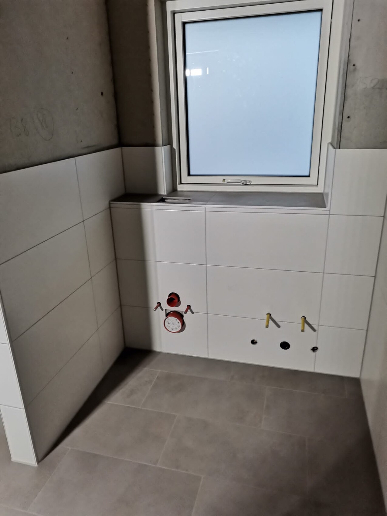 Wand mit rotem Wasserzähler, zwei gelben Armaturen, Abflussloch, Fenster über weißen Fliesen.