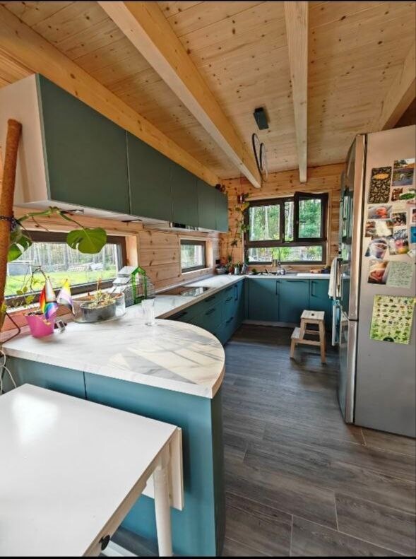 Kitchen in a wooden house with blue lower cabinets, light countertop, window front and refrigerator.