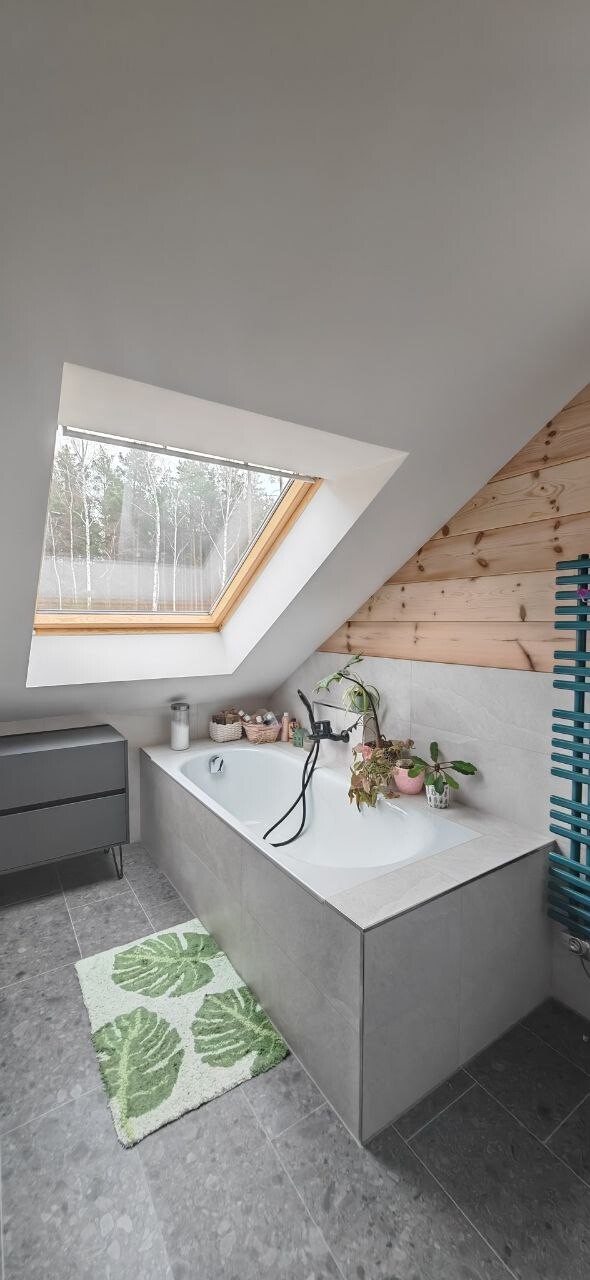 Attic bathroom with bathtub under skylight, wooden wall, plants, and green bath mat.