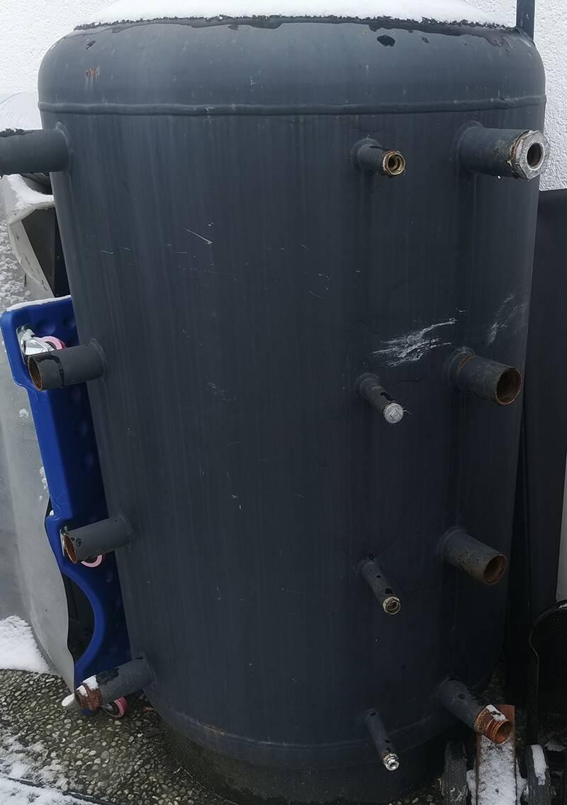 Large dark cylindrical tank with multiple pipe connections, standing outside in the snow.