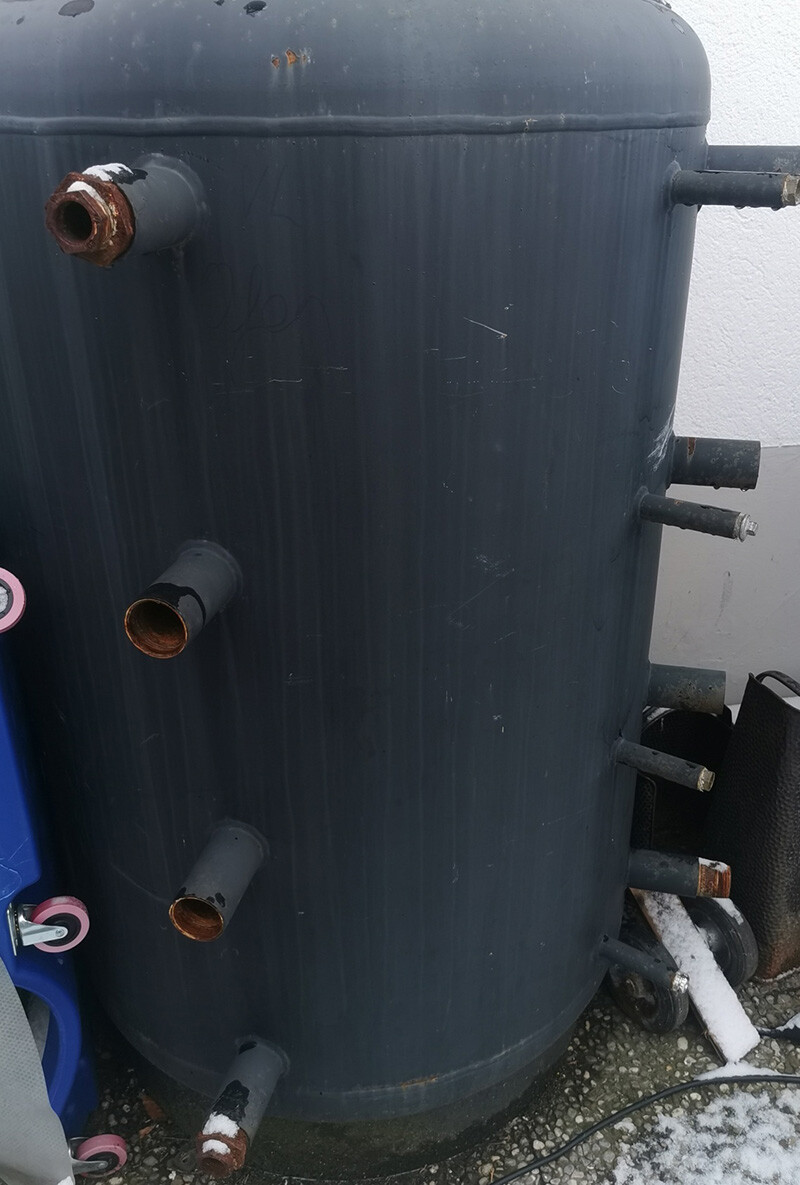 Large dark cylindrical container outdoors with several rusted pipe connections on the side.