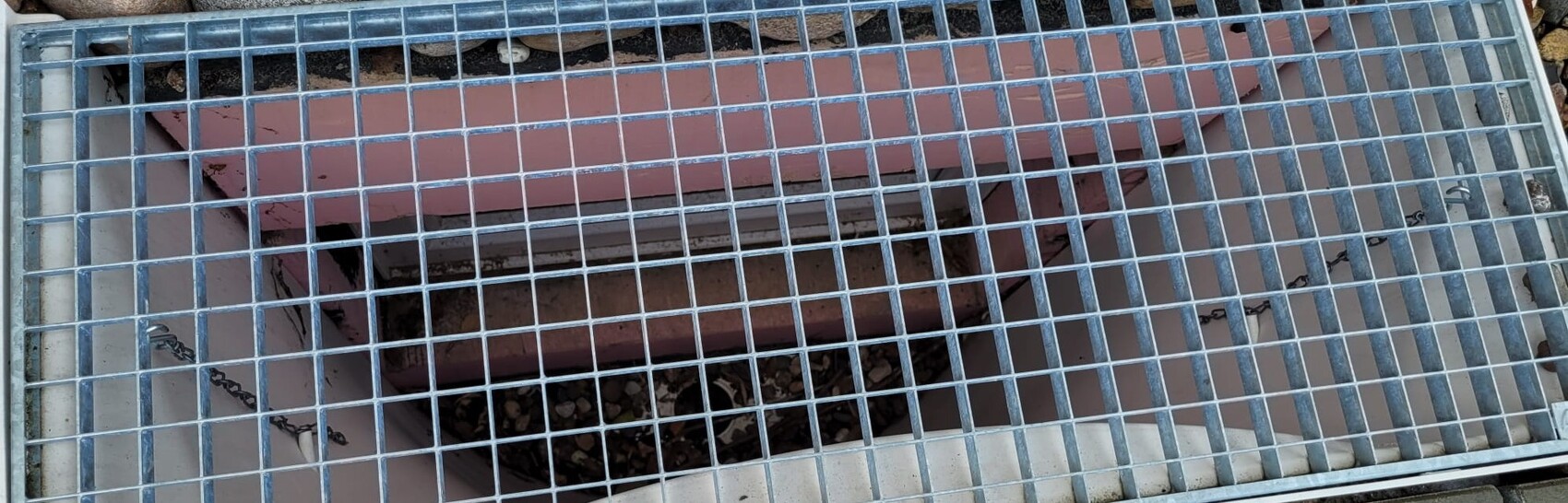 Metal grid cover over open hole with pink interior walls, gravel and dirt below.