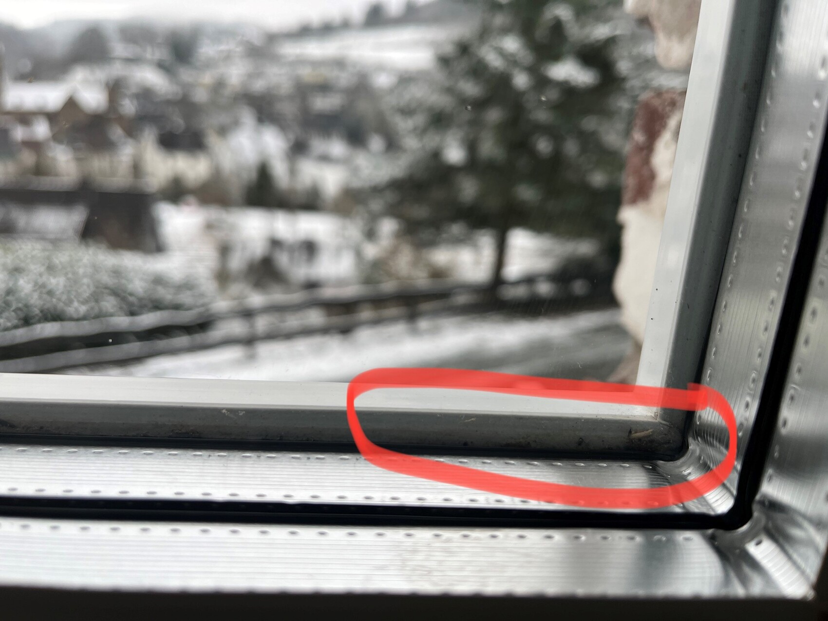 Nahaufnahme eines Metall-Fensterrahmens mit Dichtung; rote Markierung zeigt Spalt, verschneit draußen.