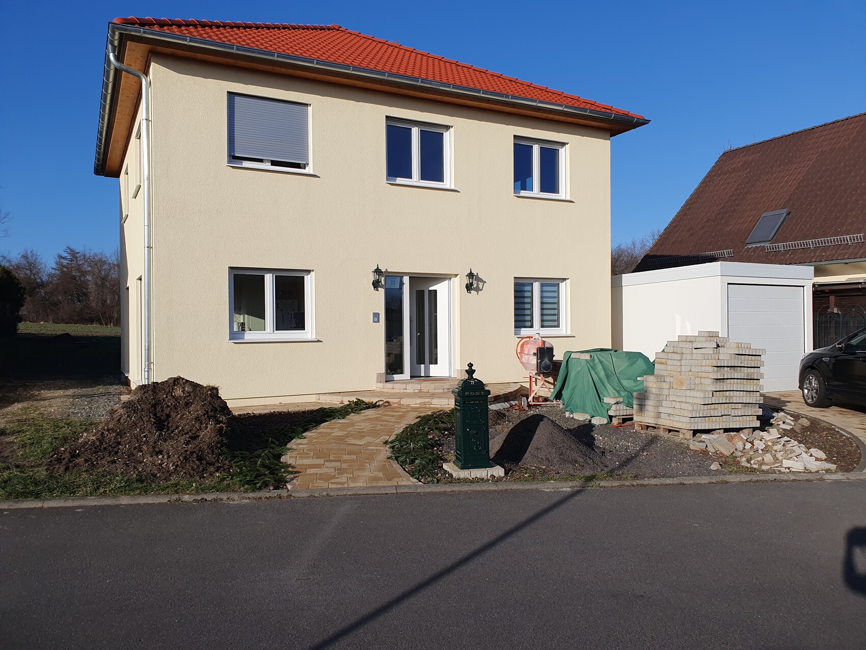 Two-story beige house with red roof; construction work at the entrance, material stacks in the garden.