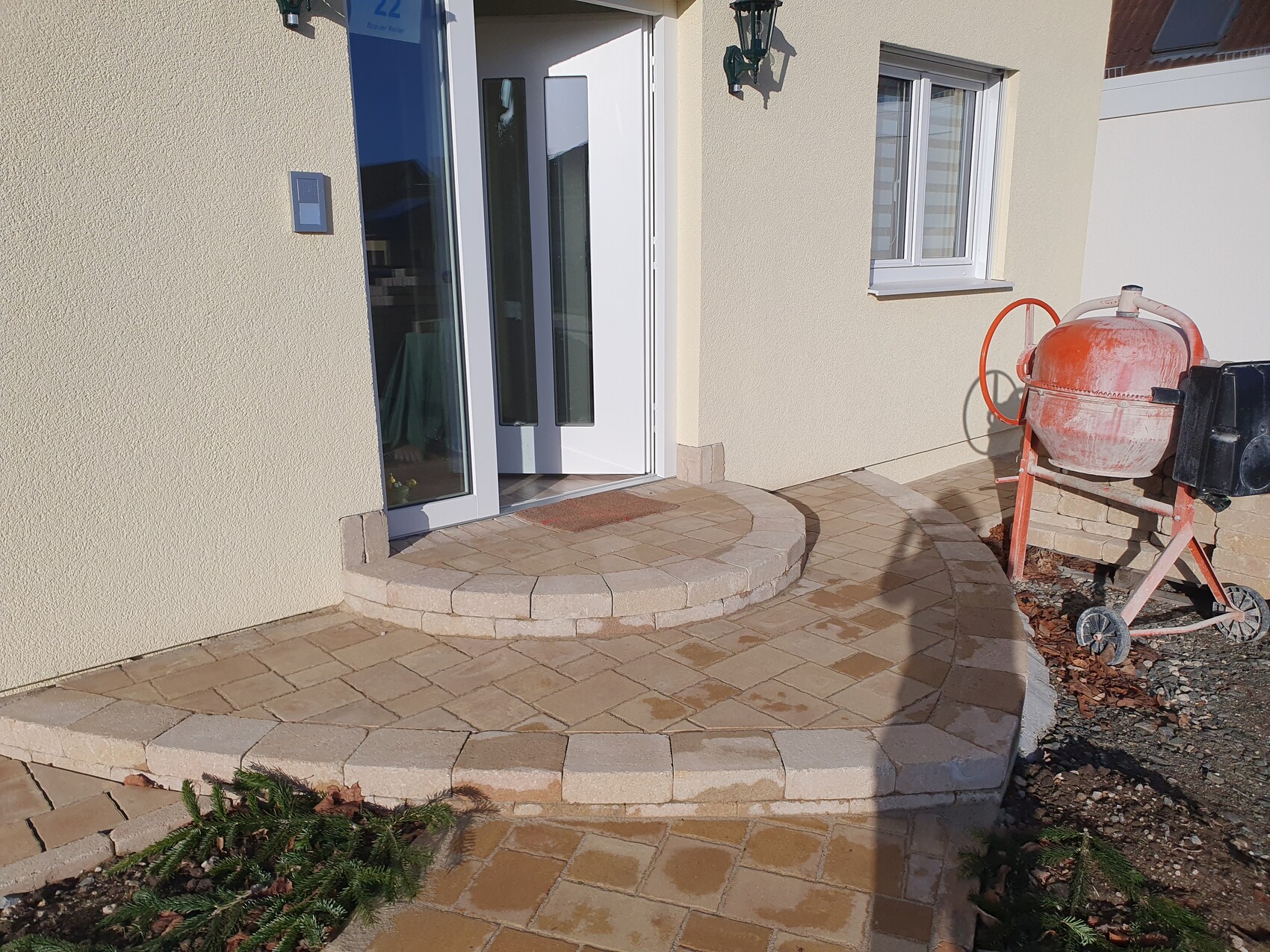 Beige facade with white glass door, circular stone slabs in front of entrance, concrete mixer on the right.
