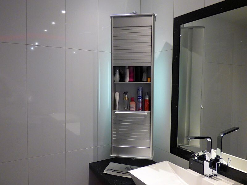 Tall, narrow metal shelf in bathroom with hygiene products in open compartments in front of mirror.