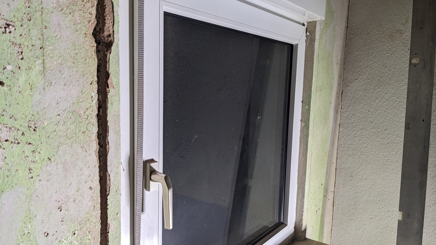 White window frame with handle in front of greenish plastered, cracked wall.