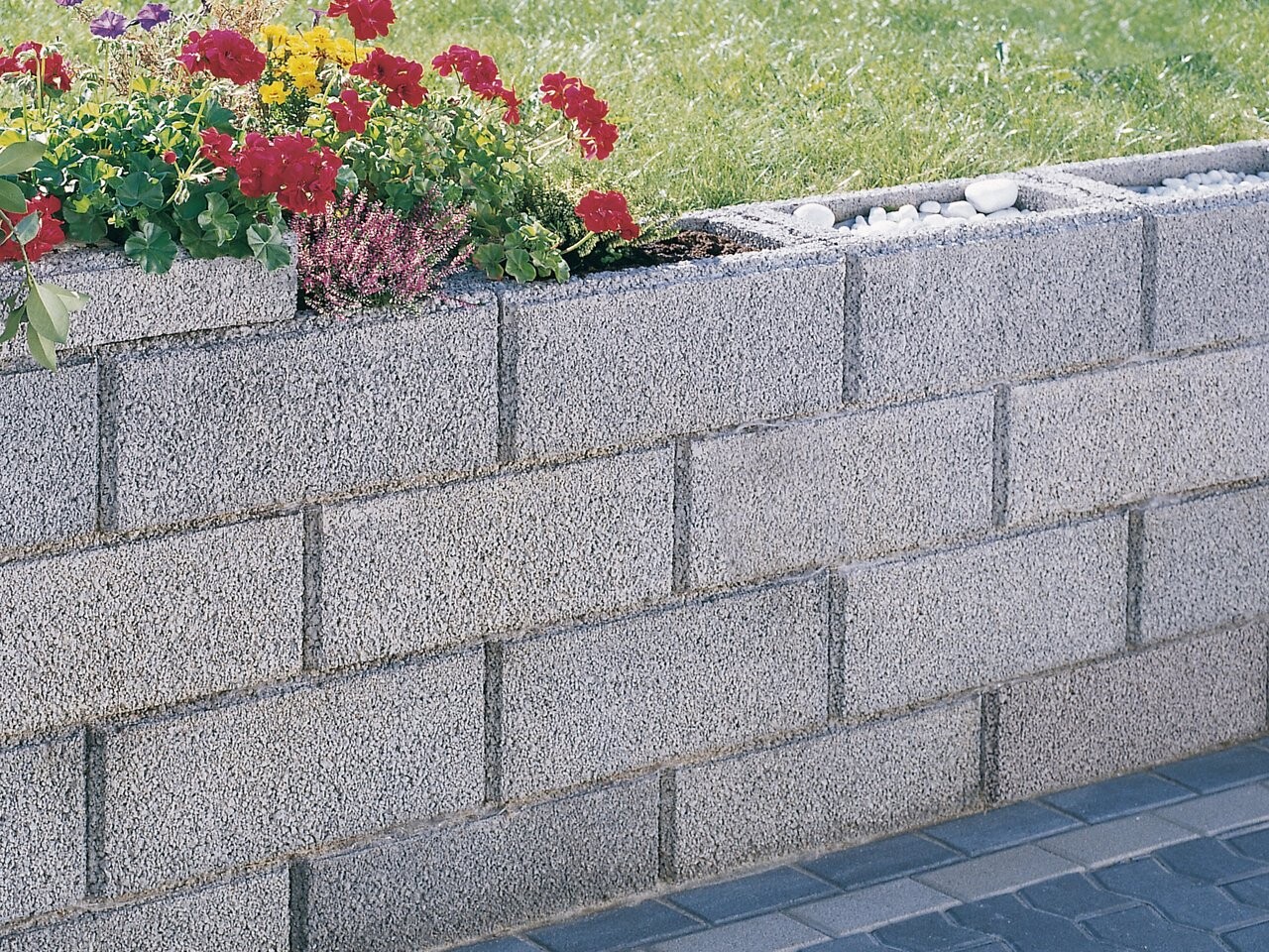 Grey block wall as a border for a flower bed with red geraniums, green foliage, and purple heather, gravel at the edge.