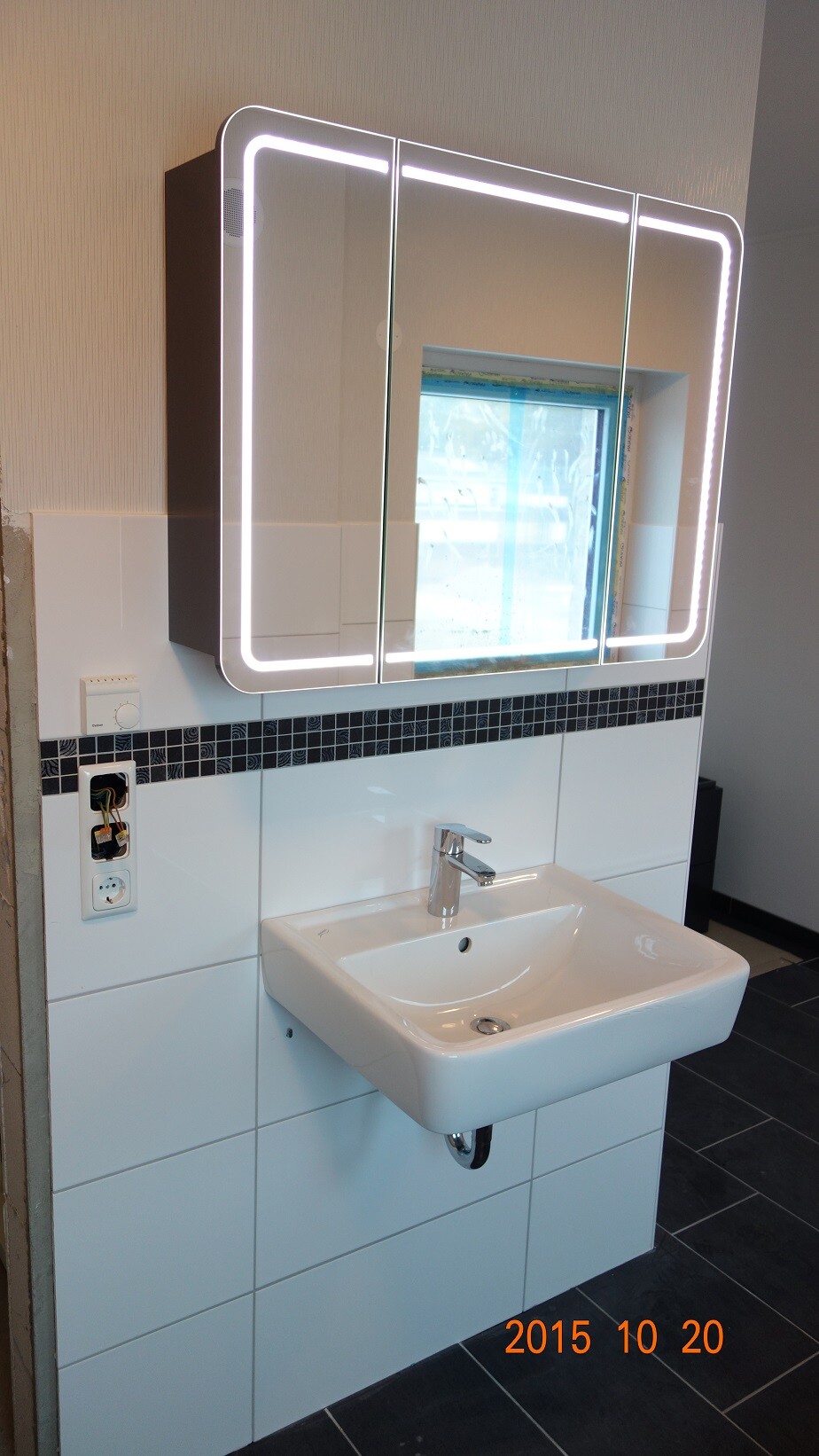 Modern LED mirror cabinet with edge lighting above a white sink on a tiled wall.