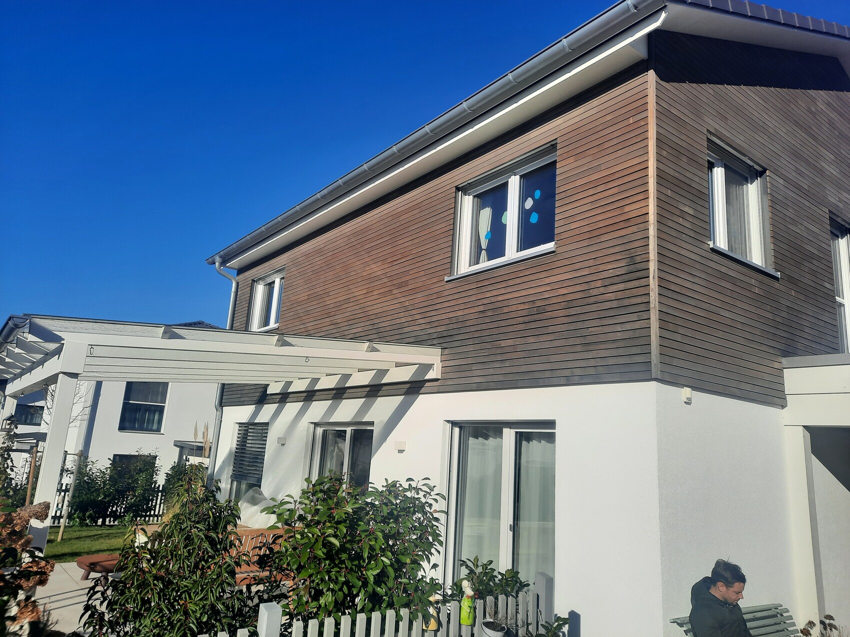 Modern house: white base, wooden cladding above, pergola and garden; person sitting on bench.