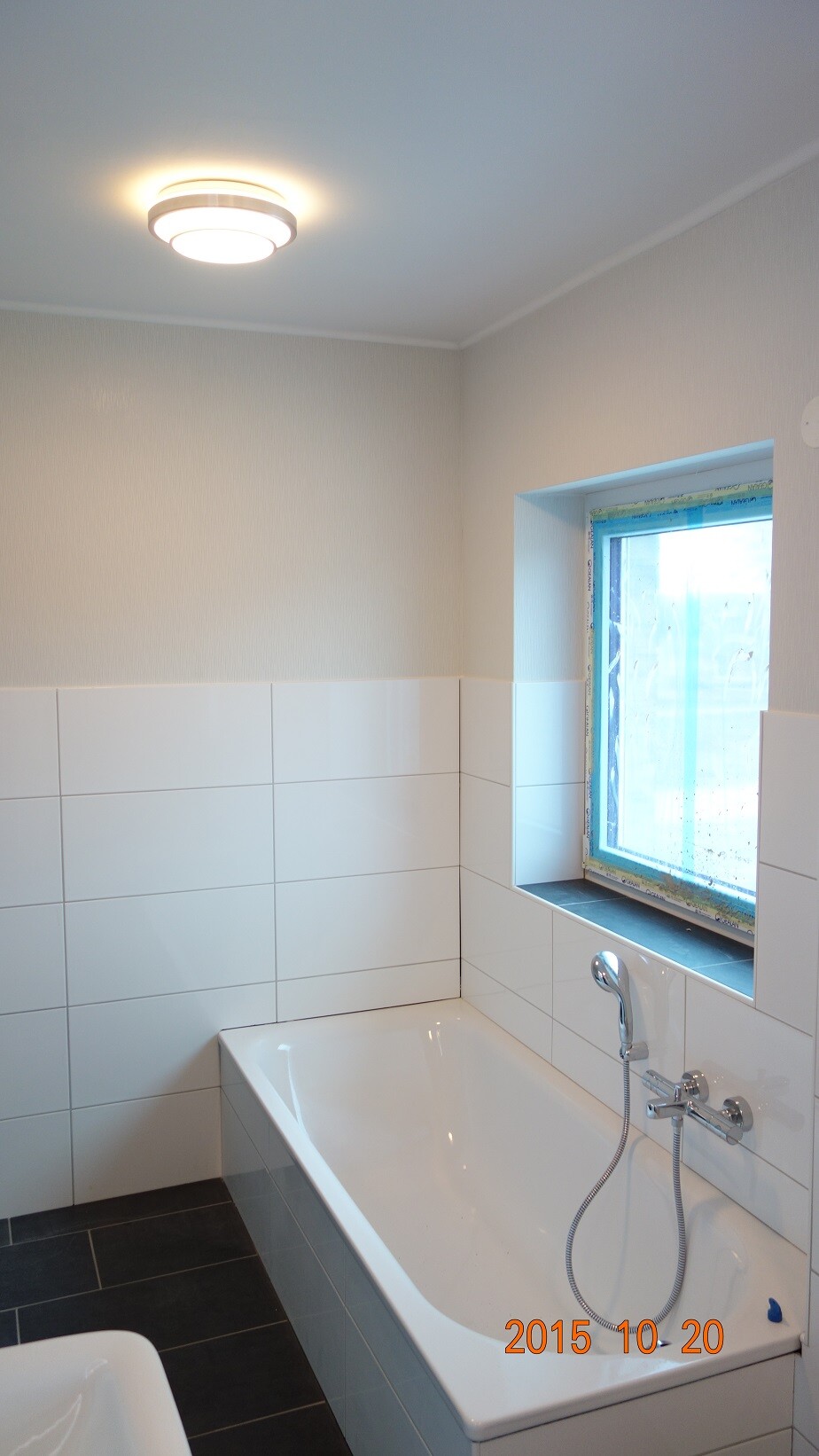 Bathroom with a white bathtub, white wall tiles, dark floor tiles, and a window on the right.