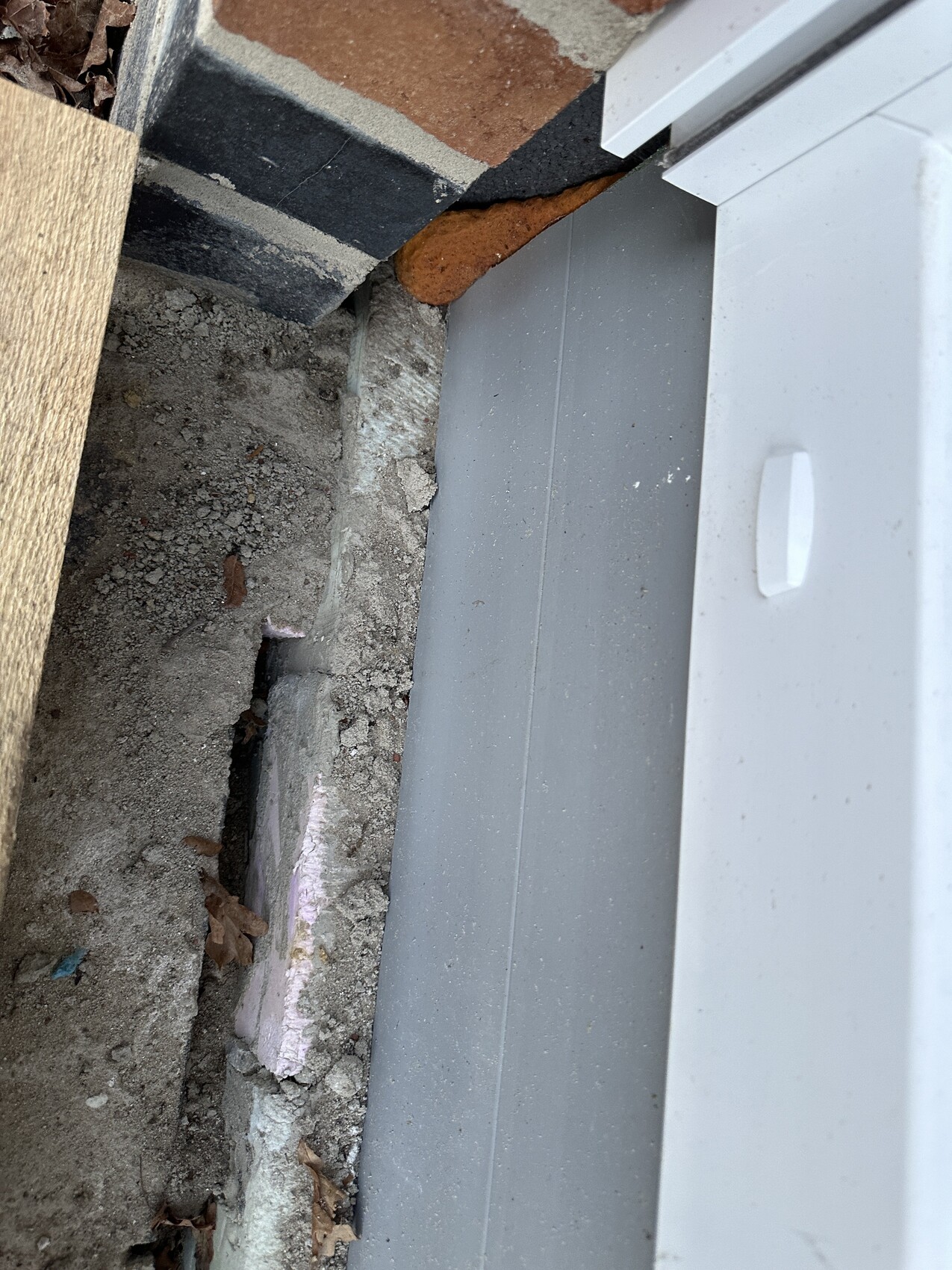 Nahaufnahme einer Baustelle: Ziegelwand, Betonfundament und grauer Tür-/Fensterrahmen mit Griff.