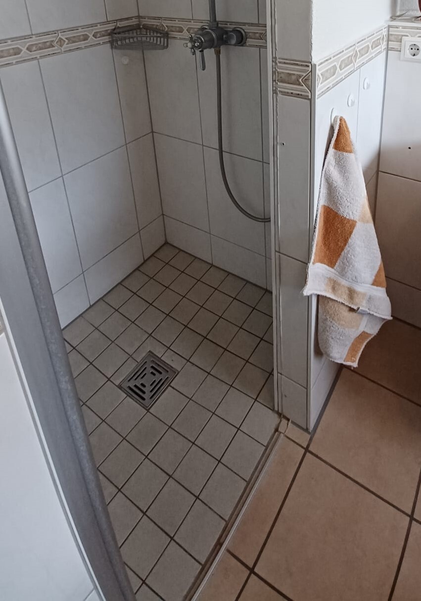 Small shower bathroom with wall-mounted handheld shower, tiled floor, floor drain, orange-white patterned towel.