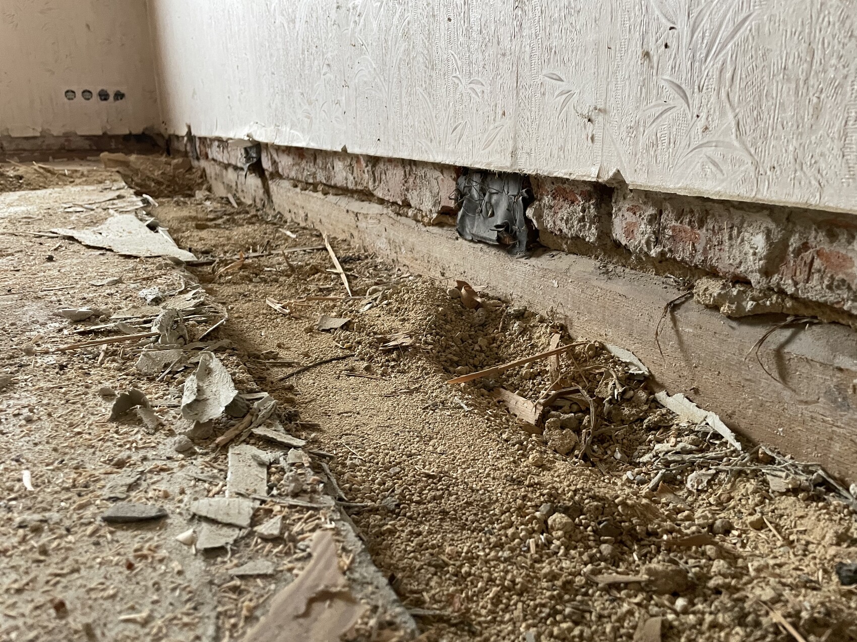 Basement room with damaged wall covering, exposed bricks, and floor made of soil and debris.