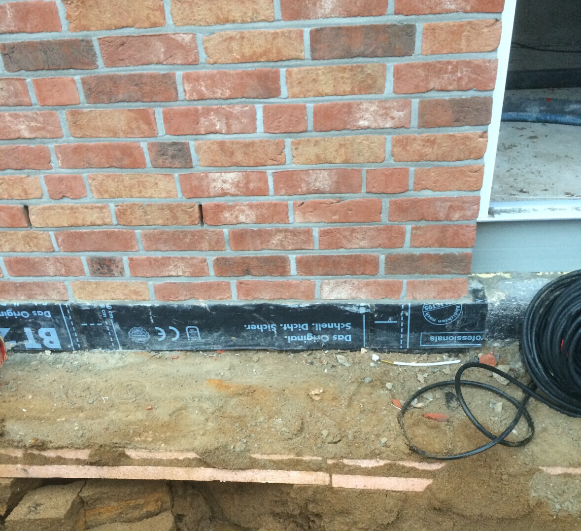 Exterior of a brick house with black foundation waterproofing and a cable reel on the ground.