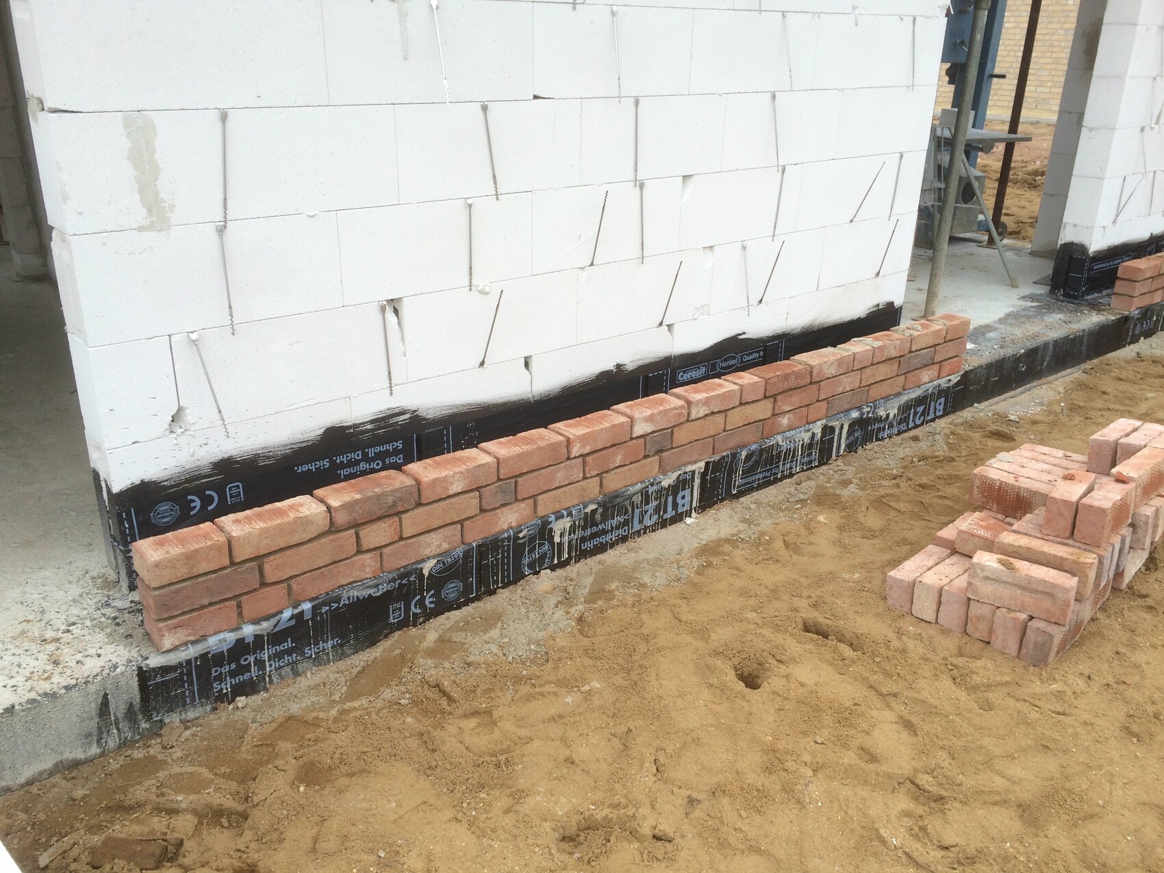 Building wall made of white aerated concrete blocks with black waterproofing, in front a row of bricks is being laid.