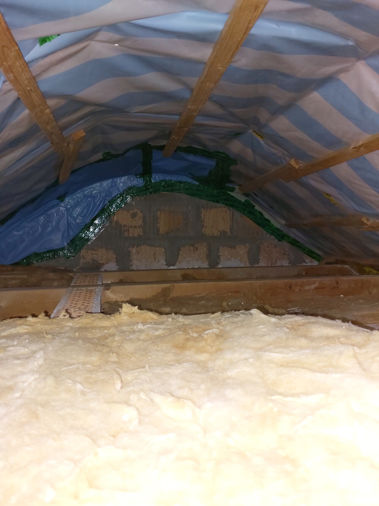 Attic with insulation, wooden beams, blue-white foil, green tape around arch, brick wall.