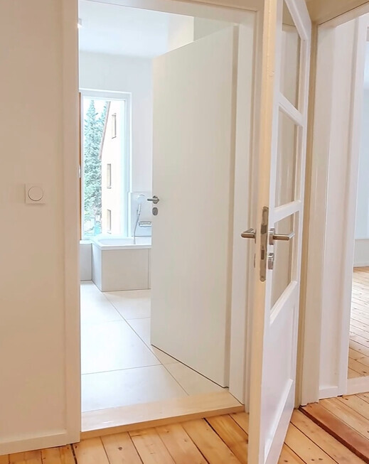 Open door in the hallway leading to a bright bathroom with a white bathtub by the window.