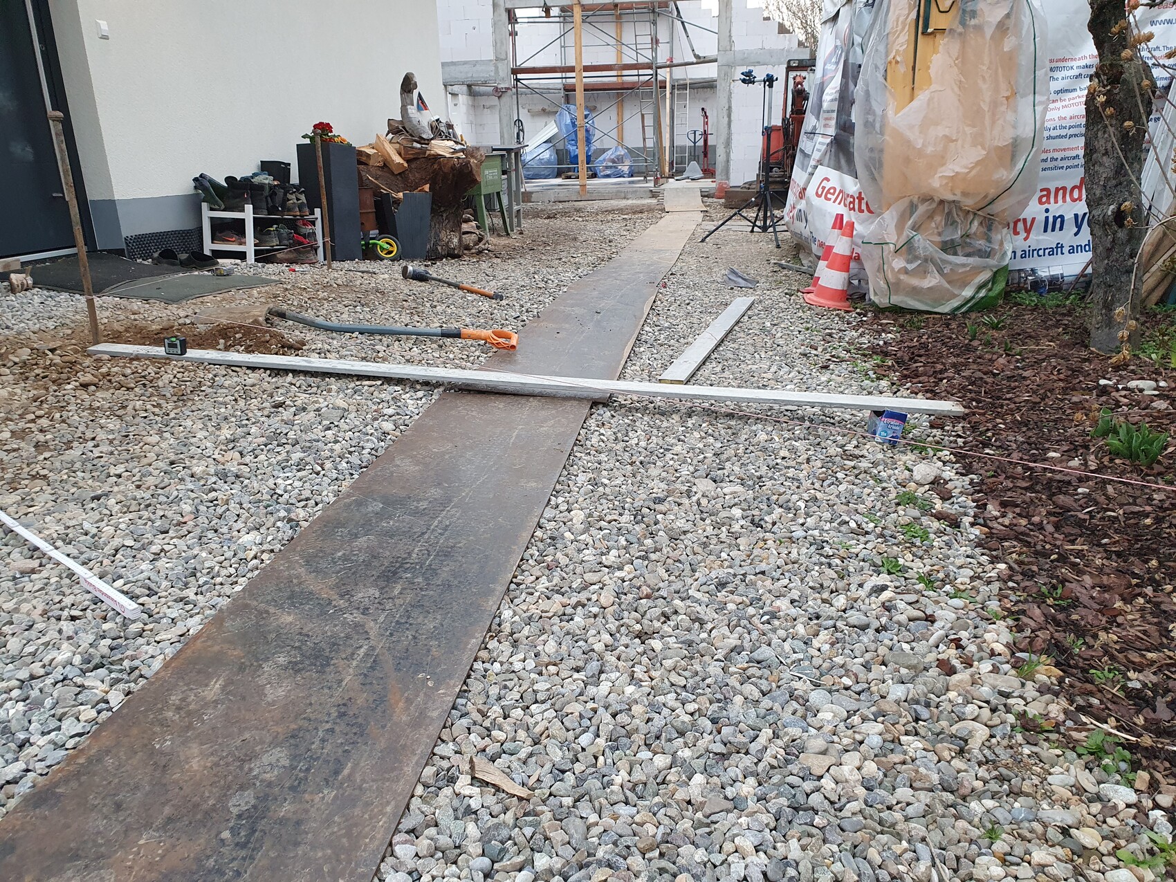 Außenbaustelle mit provisorischem Holzweg, Bauwerkzeugen, Kies und Planen.