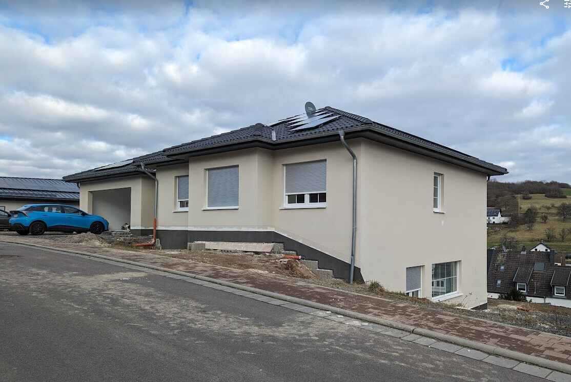Wohnhaus am Hang mit Solarmodulen, blauem Auto vor Garage, wolkiger Himmel.