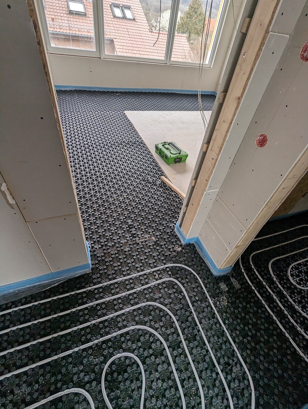 Raum im Bau mit Fußbodenheizung: schwarze Platten, weiße Rohre, grüne Werkzeugbox.