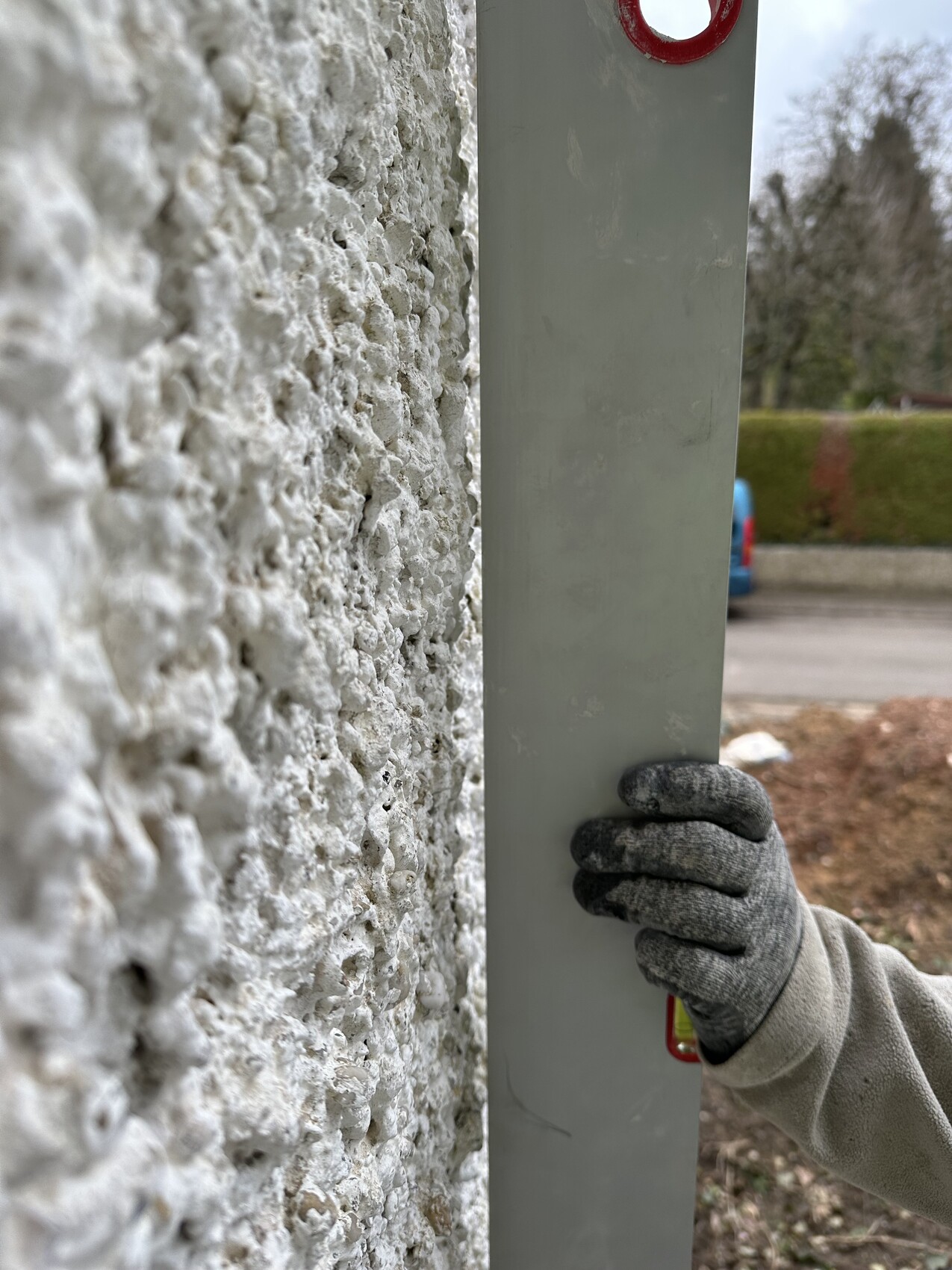 Hand in graue Arbeitshandschuh hält eine graue Metallstange an einer rauen weißen Wand.