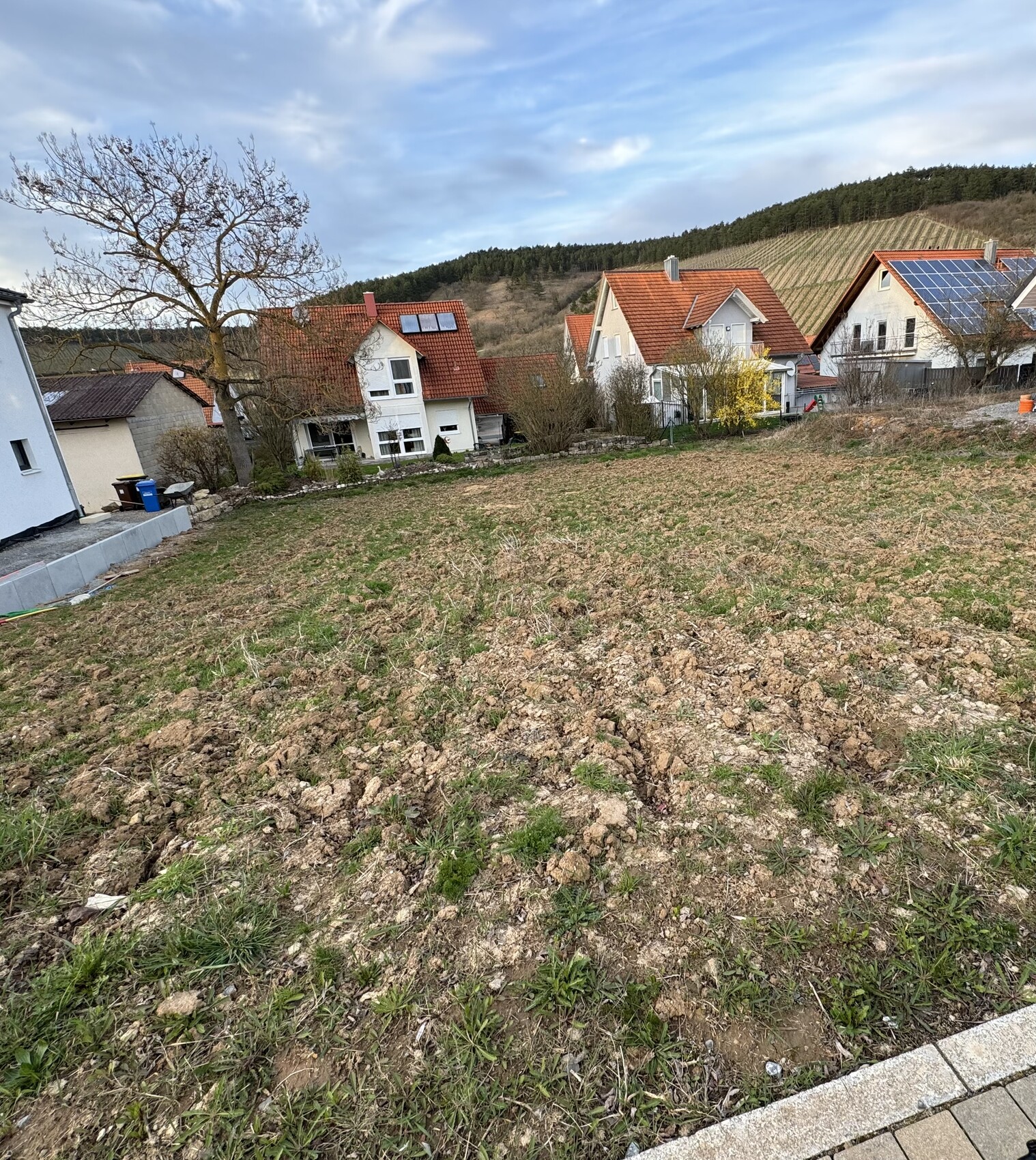 Freier Baugrund vor Wohnhäusern mit roten Dächern, Kiesboden und Hügel im Hintergrund.
