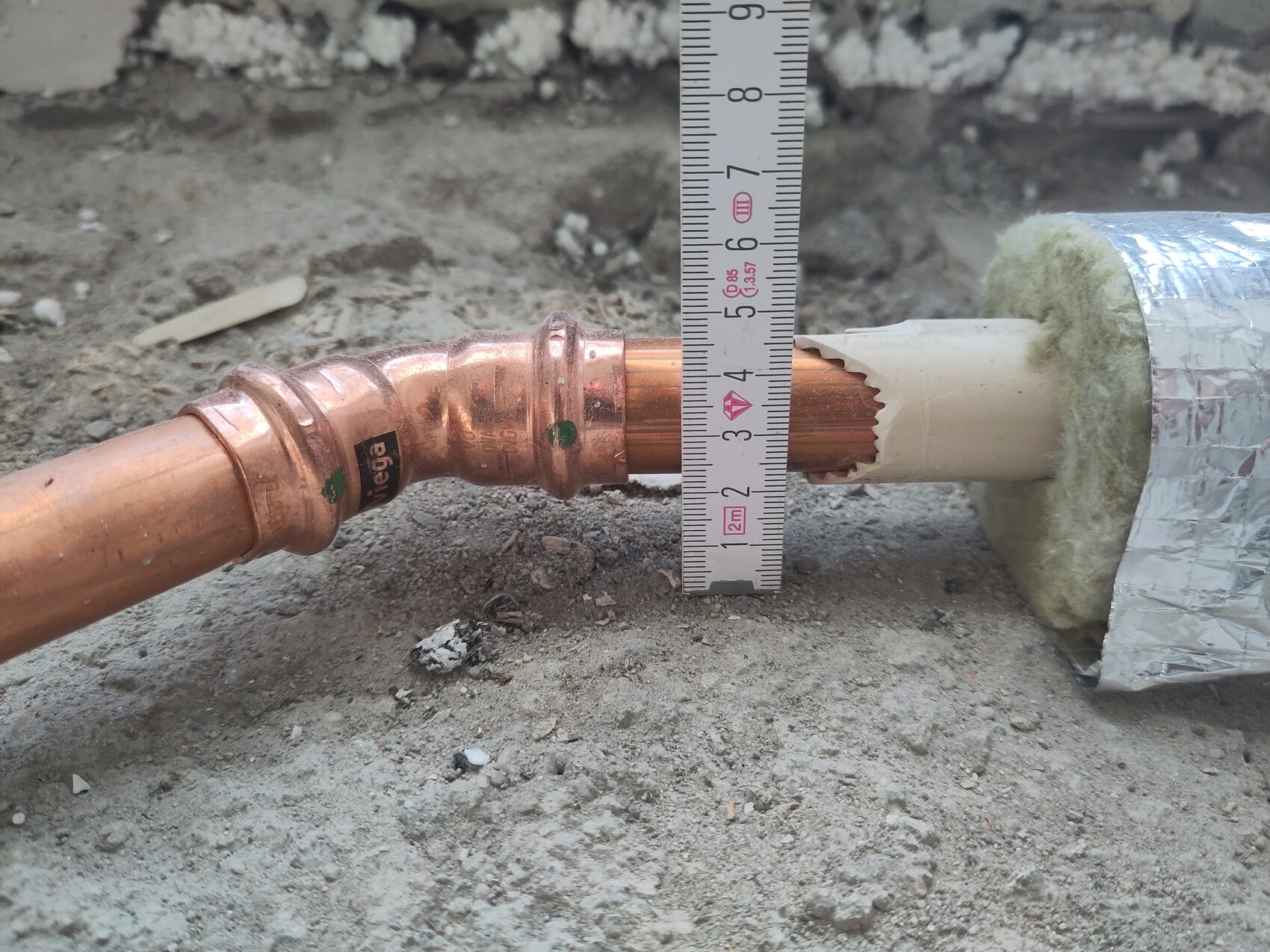 Copper pipe with fitting, measuring tape next to it; insulated pipe passing through a wall opening on a construction site.