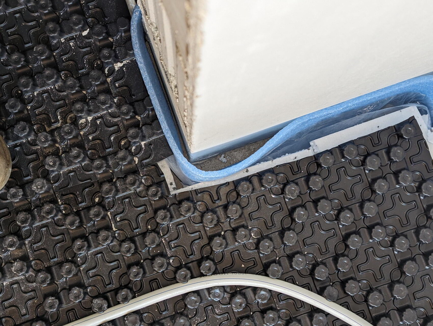 Black studded installation panel for underfloor heating, blue insulation on the wall, white pipe.