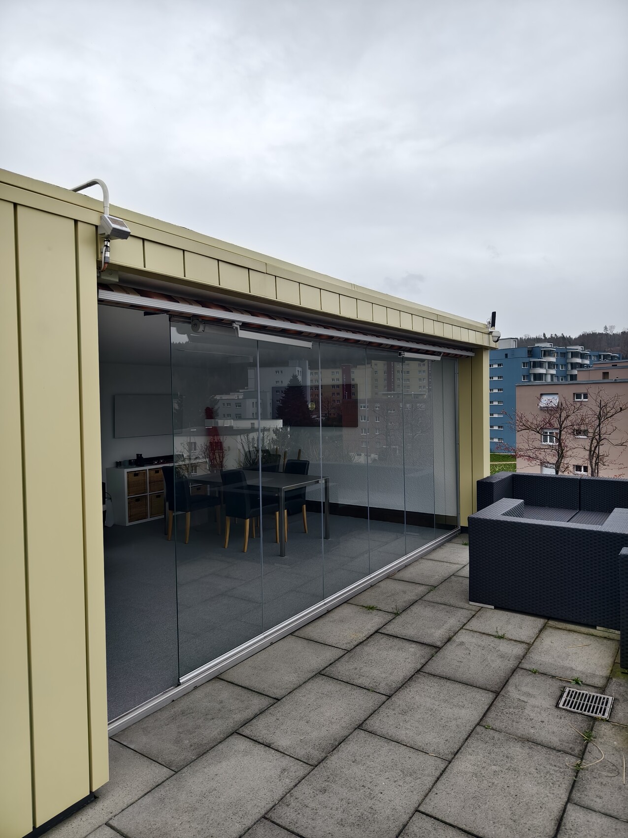 Auf einer Dachterrasse mit Glasfront; Innenraum mit Esstisch und Stühlen sichtbar.