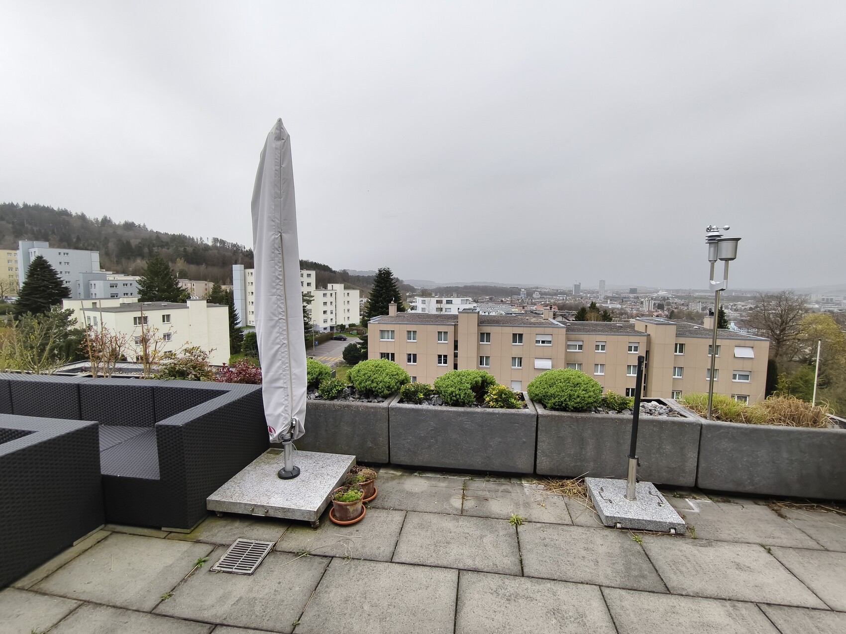 Dachterasse mit schwarzen Lounge-Möbeln, geschlossenem Sonnenschirm, Pflanzkübeln und Stadtblick.