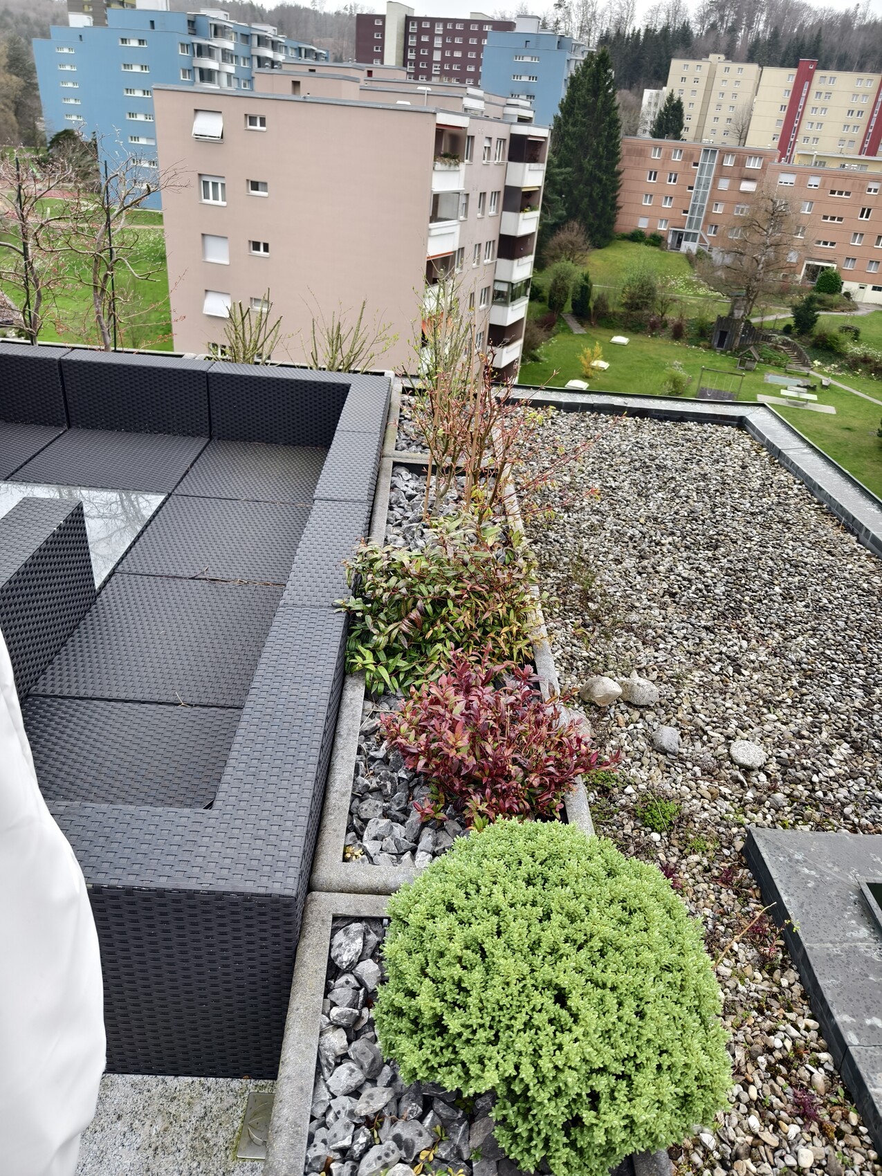 Dachterasse mit dunkler Lounge aus Rattan, Pflanzkübeln und Kies, Blick auf bunte Wohnblocks.